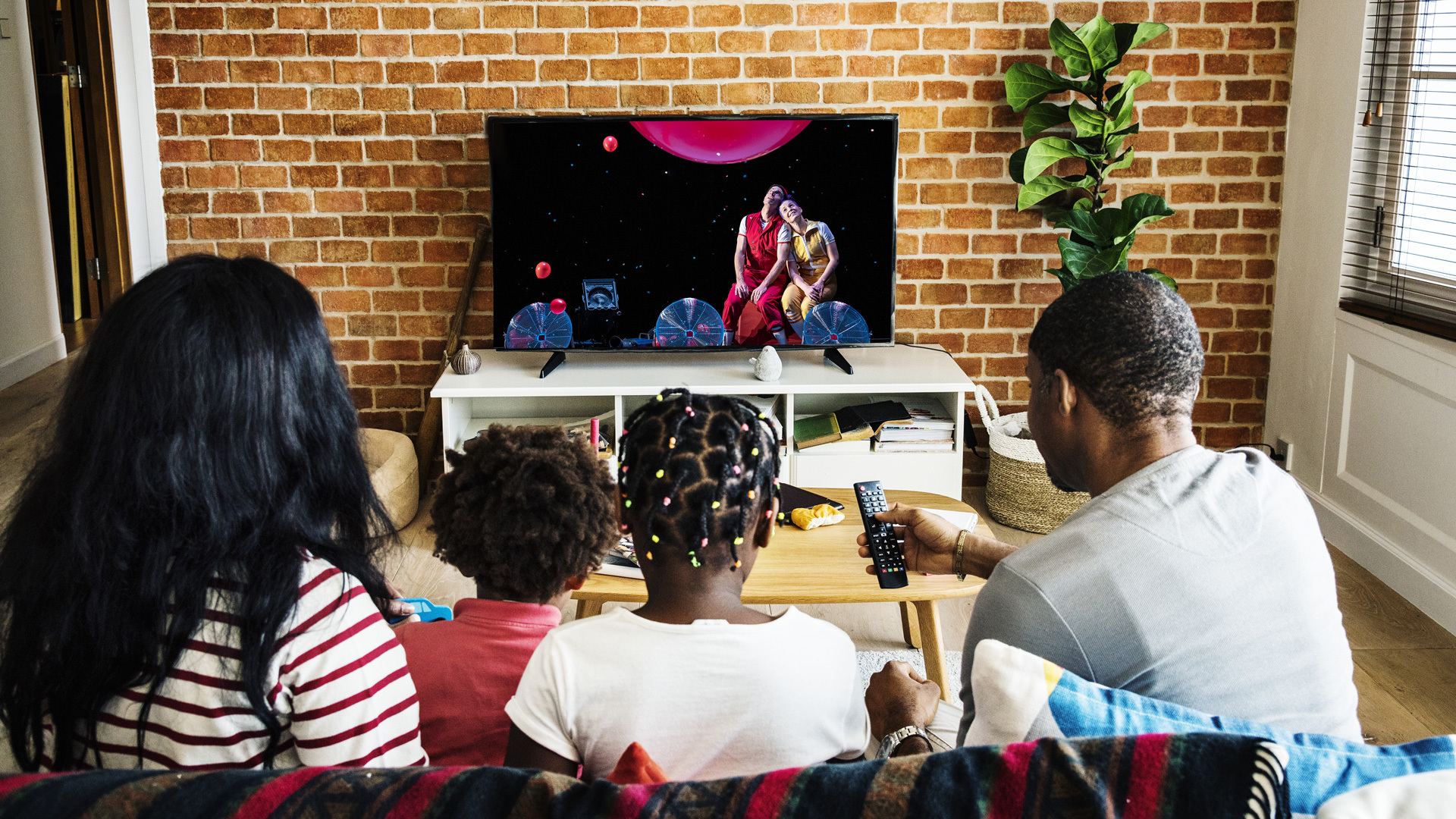 Two parents and two kids sit on a couch watching a performance of Air Play stream on their TV.