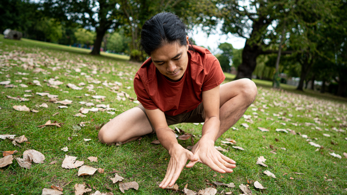 A man, crouched down on the grass, puts his hands together in the shape of a butterfly.