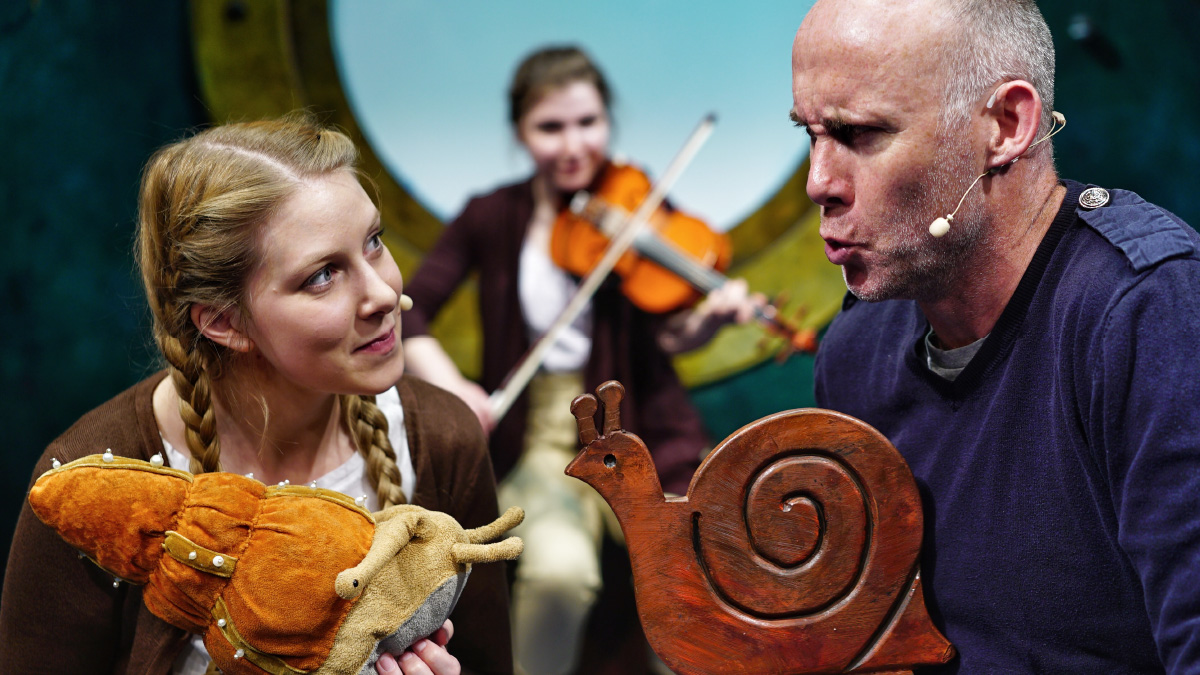 Two members of the cast of The Snail and the Whale in a scene holding snail puppets. Behind them is a musician playing the violin.