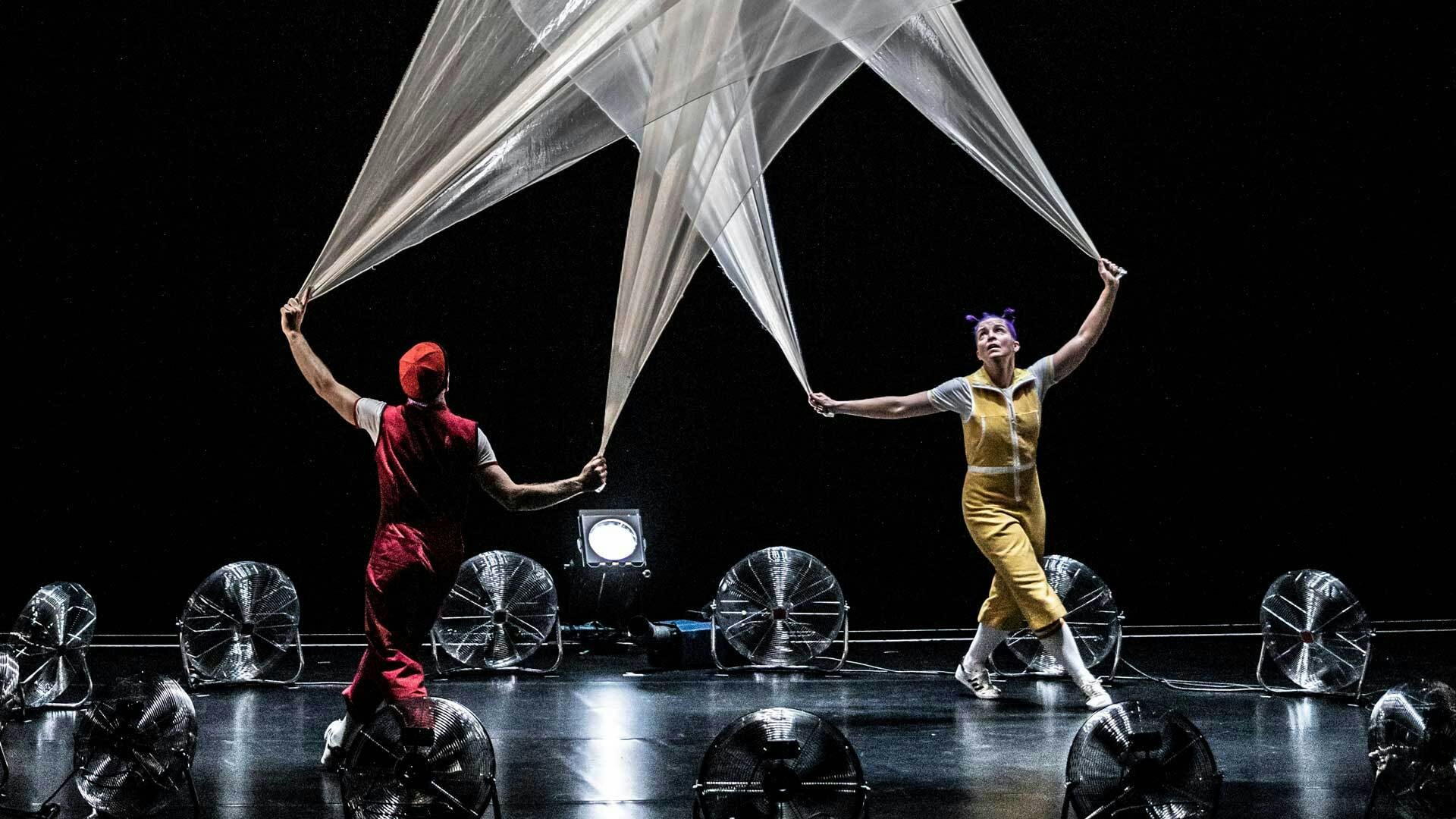 Acrobuffos hold white fabric silks above their heads as it swirls in the wind created by a circle of fans.