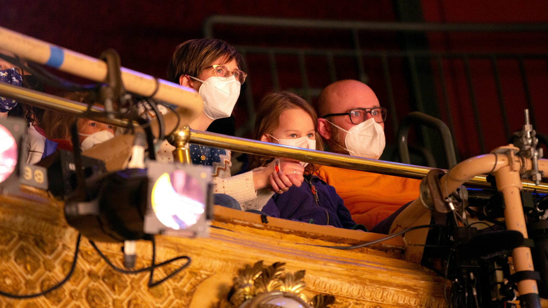 A masked family with two adults and one kid watch a show from seats in the balcony.