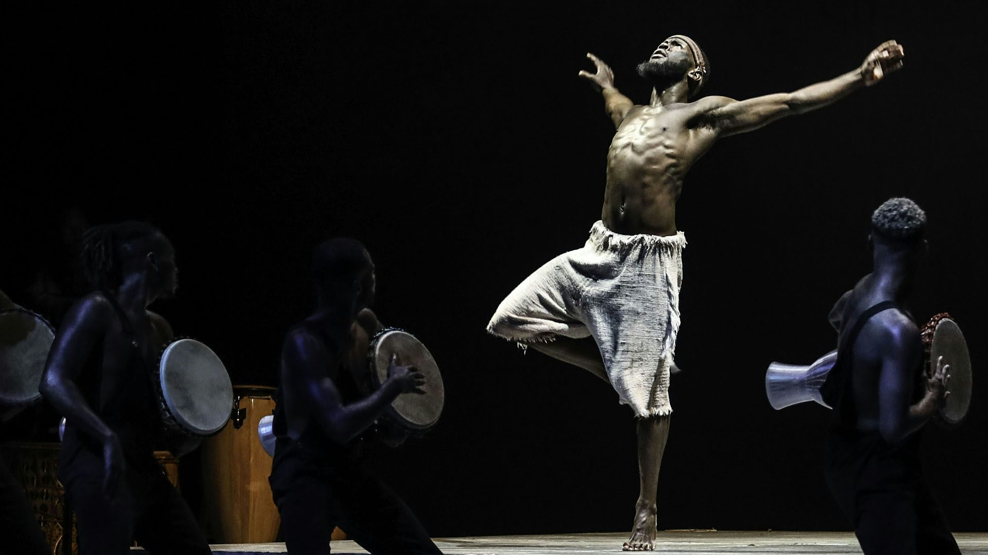A male solo dancer, stands on one foot, looking up at the sky with his arms outstretched. Around him there are three people playing hand drums.
