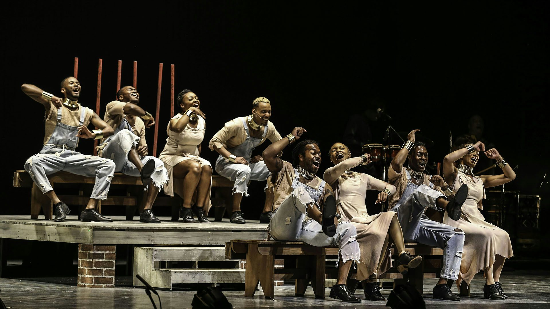 A group of dancers from Step Afrika! sit on benches in a number from Stono.