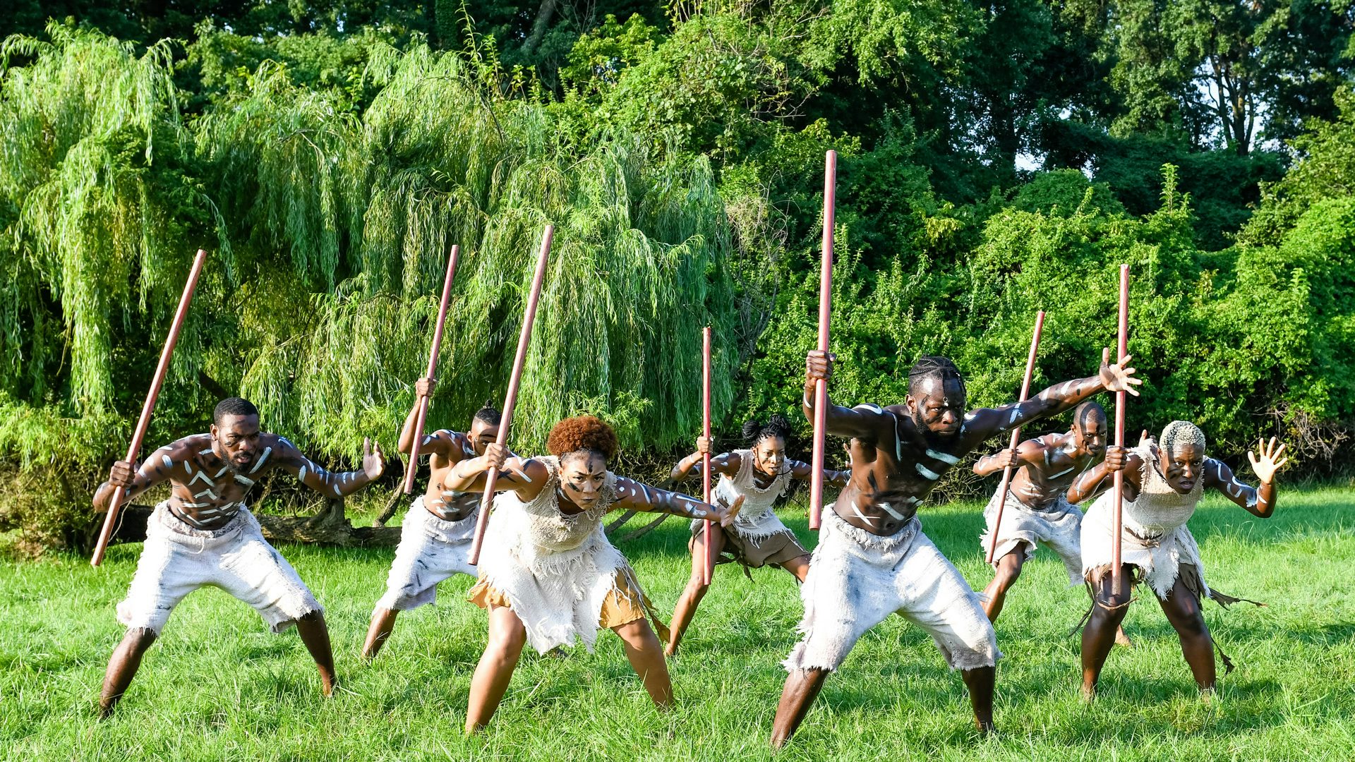 A group of dancers with African tribal body paint stand in formation, holding martial sticks, as if ready to fight.