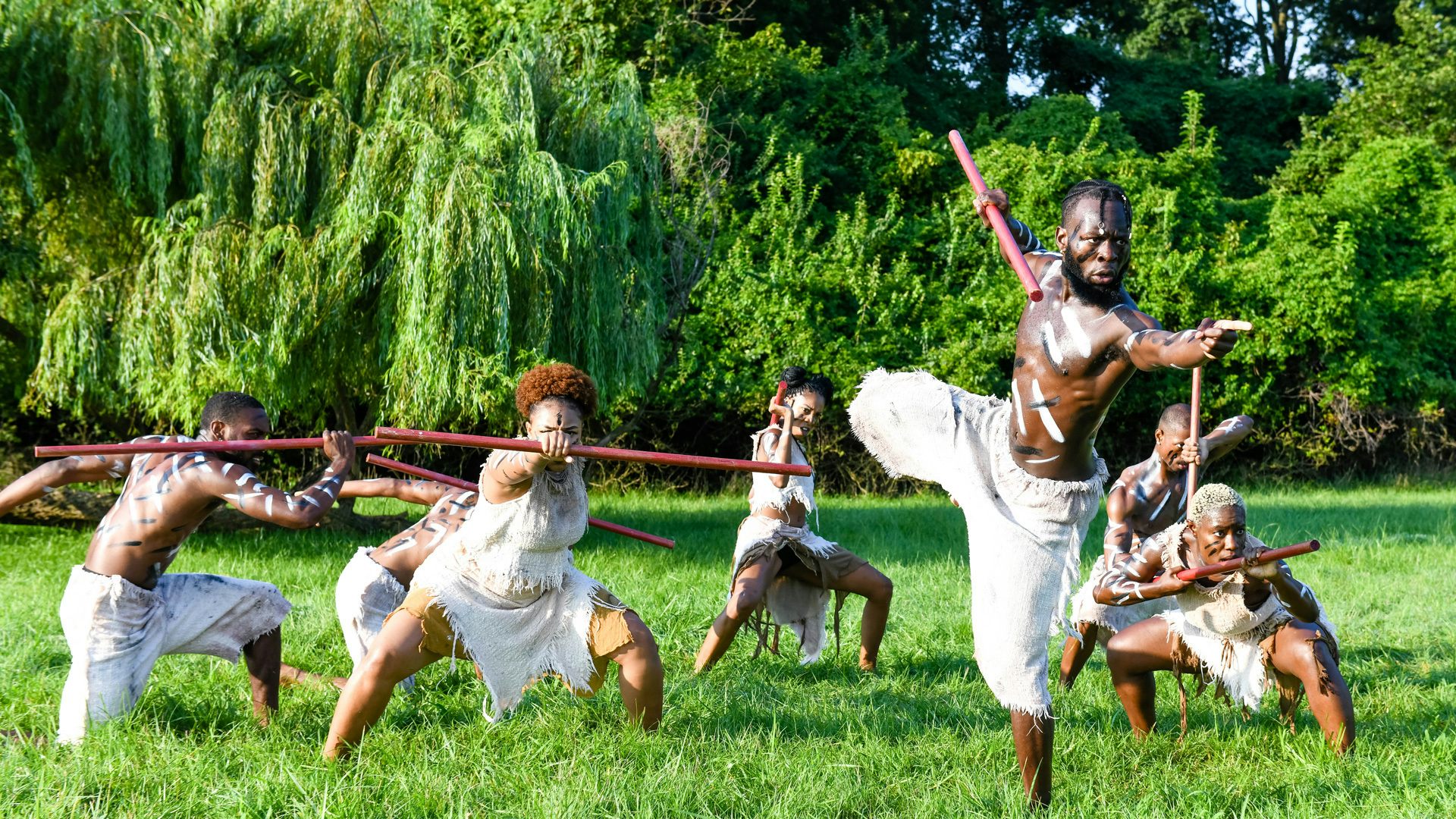 A male dancer holding a martial stick stands on one pointing. Behind, a group of six dancers crouch low to the grass.