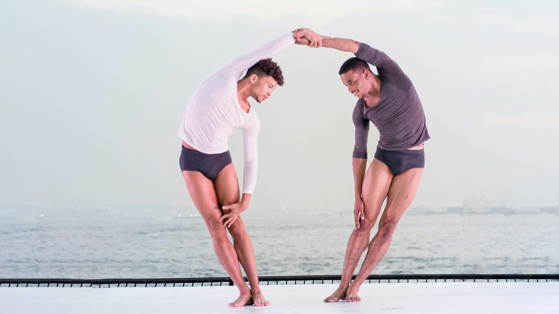 Holding each other's forearm, two male dancers lean away from each other, with curved bodies.