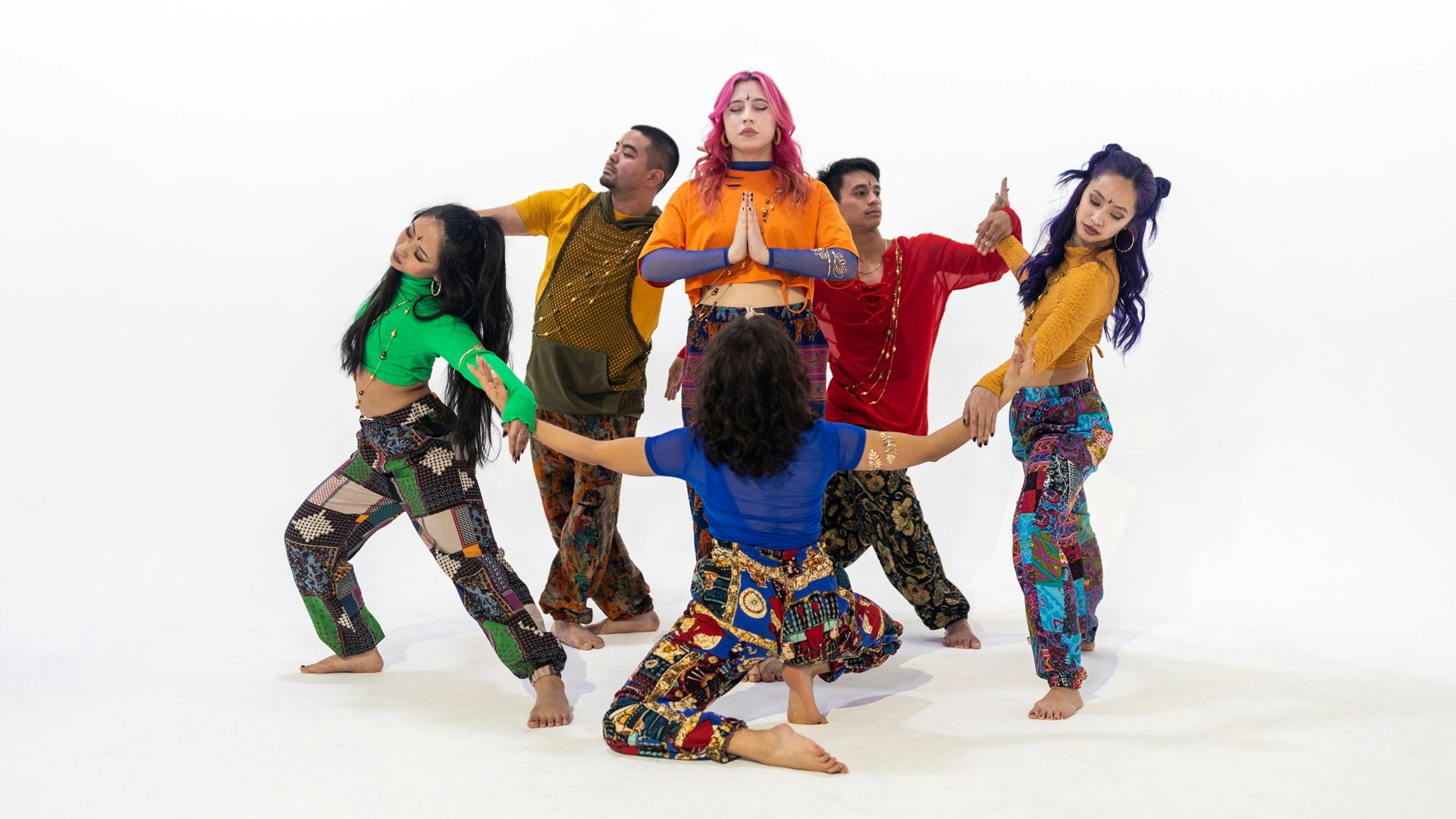 A group of dancers in colorful patterned clothes form a circle around a single dancer, standing with their eyes closed and hands in a prayer position.