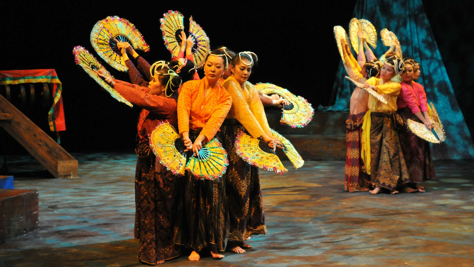 Two groups of dancers, dressed in baro't saya (traditional Filipino attire), stand back-to-back in circles, with their arms outstretched holding fans.