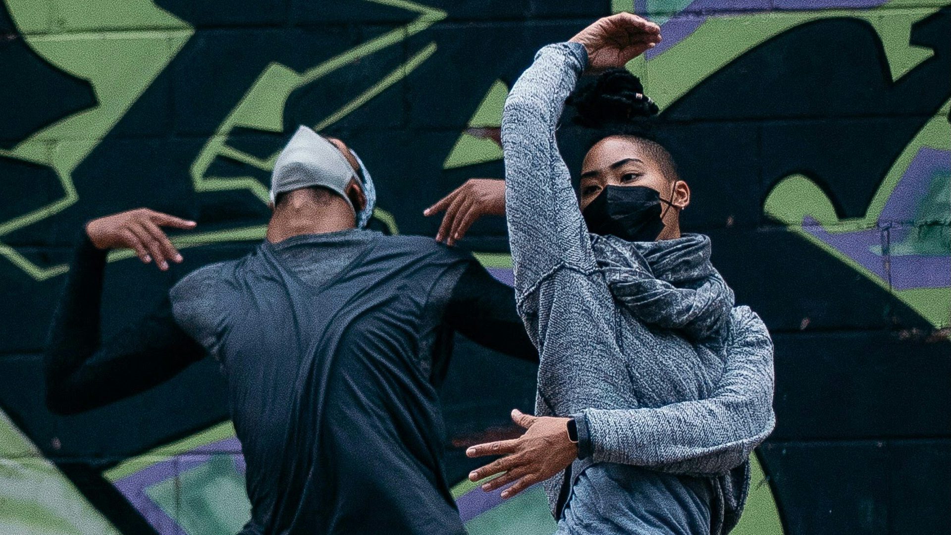 Two dancers in front of a graffitied wall. One looks up with his chest out and hands atop his shoulders. Beside him, a woman has one arm over her head and one across her body.