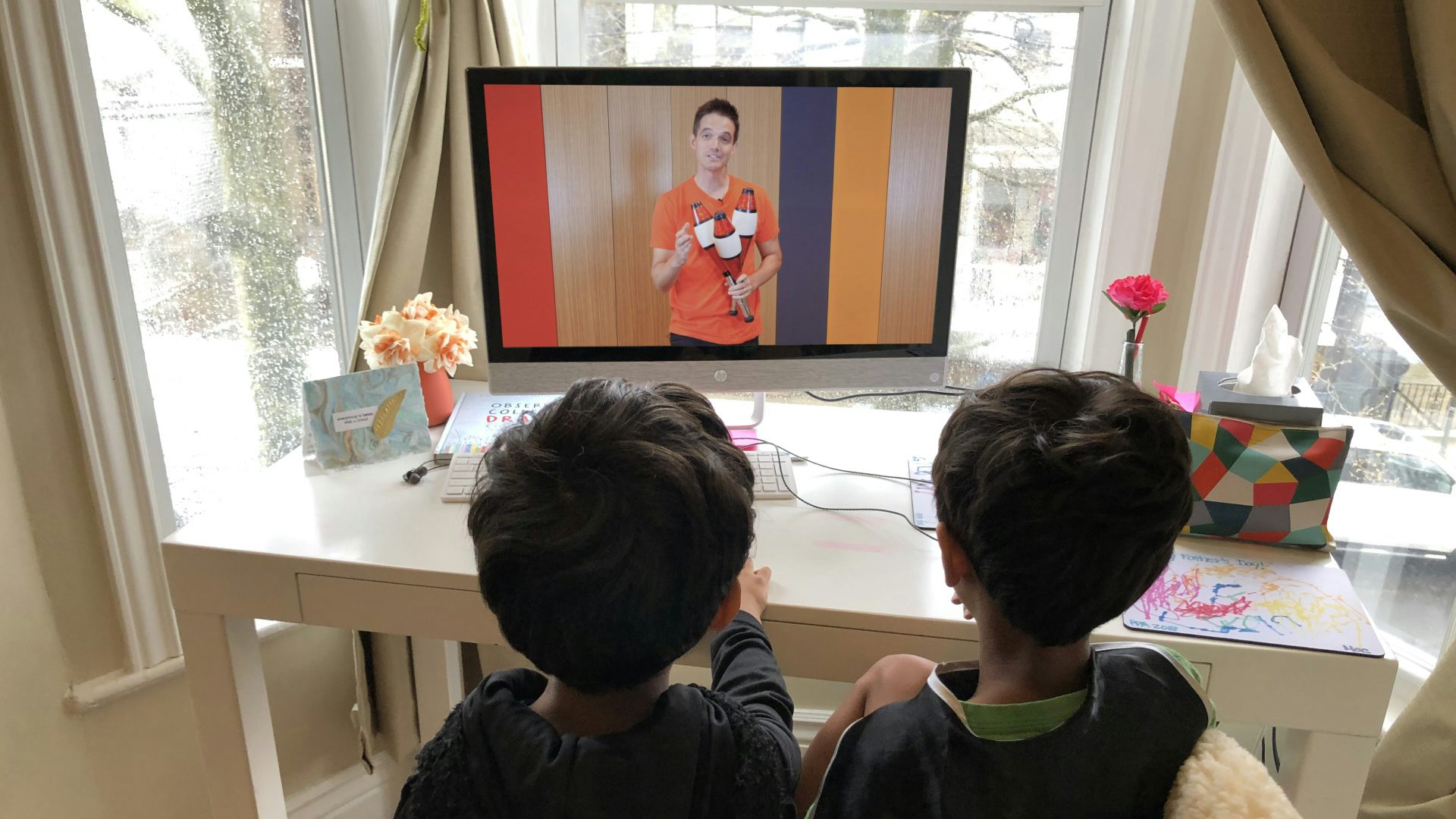 Two kids shown from behind in an at-home office as they watch a person on a computer screen speaking while holding three juggling pins