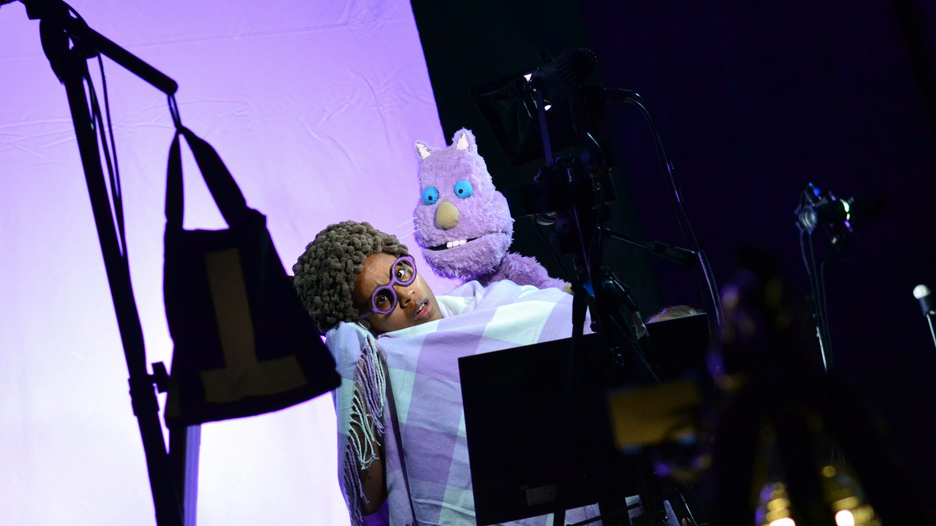 Kerry and Frankkenthaler, a purple hand puppet, look at a camera.