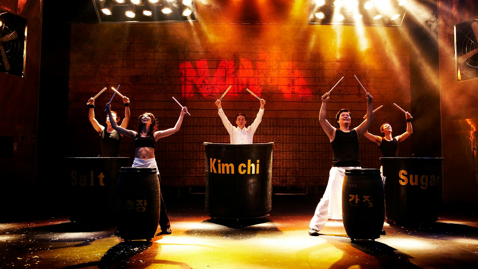 On a stage, five people stand in front of differently sized barrel drums. Each of them holds a pair of drumsticks crossed above their head.