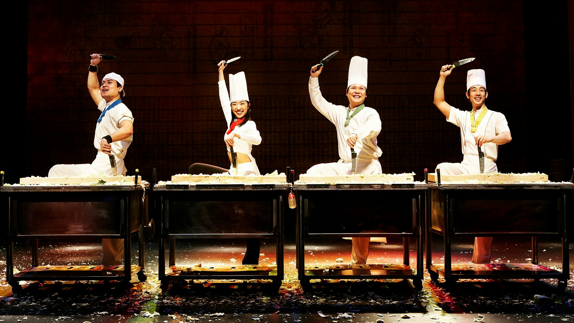 Four chefs in a line, hold a knife above their head, in a triumphant pose.