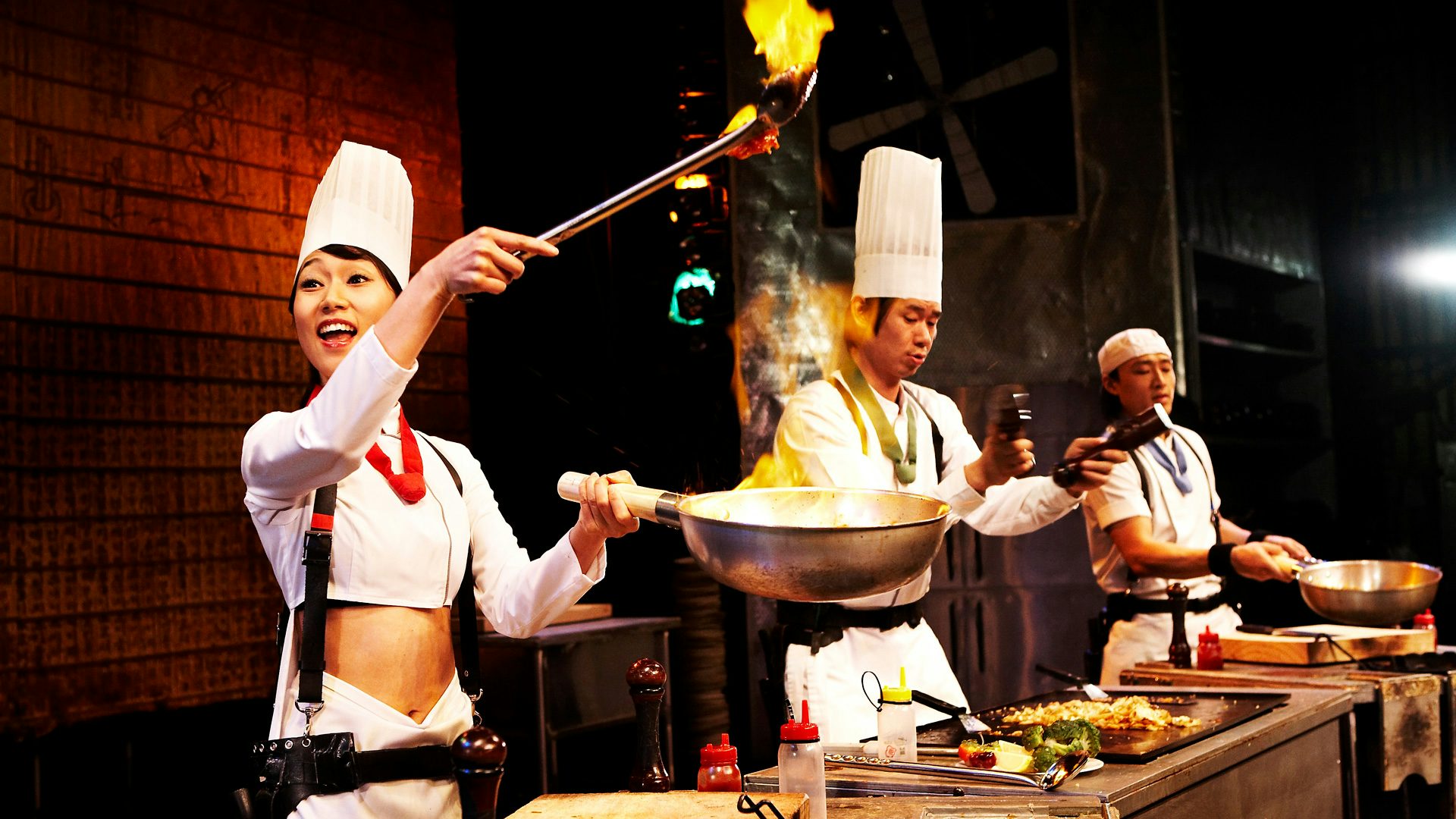 A woman, in a chef uniform, holds a flaming pair of tongs and pan.