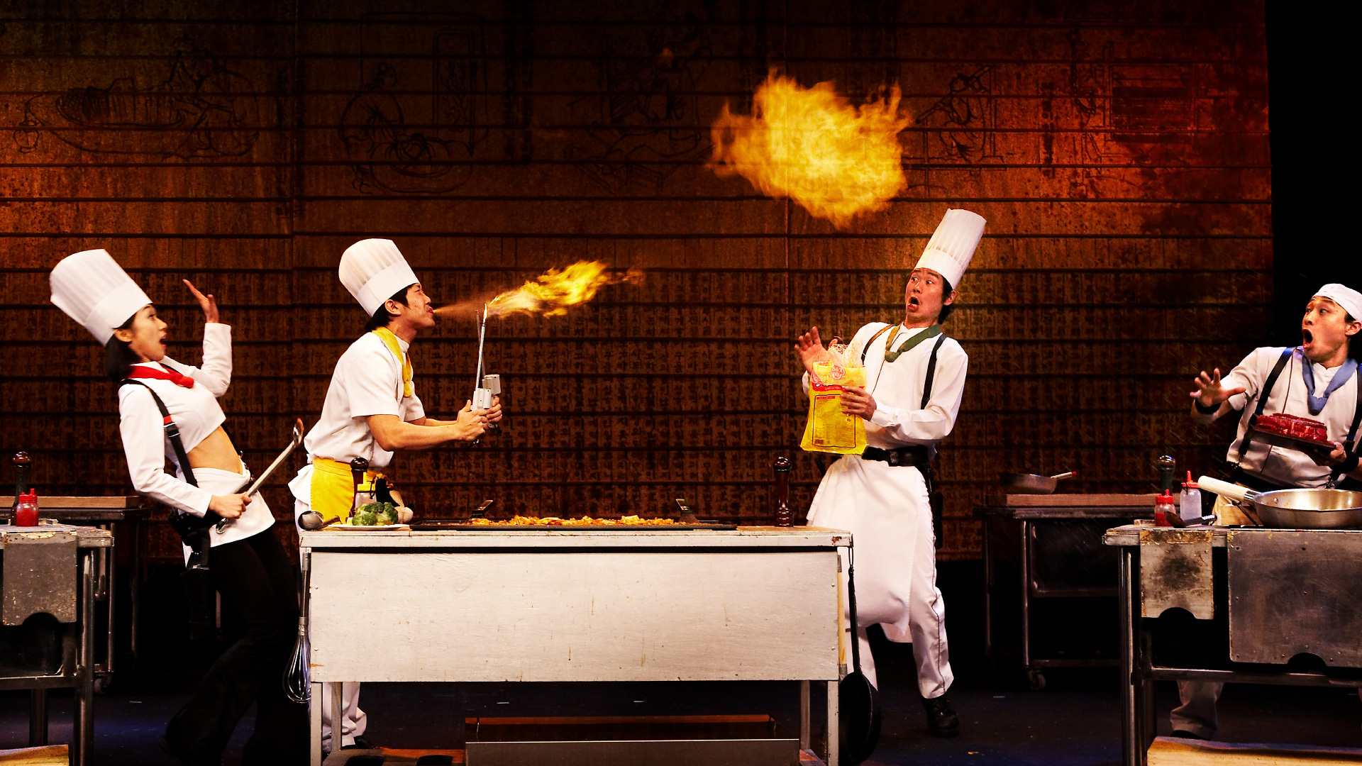 A chef breathes a cloud of fire; beside him, three surprised chefs recoil.