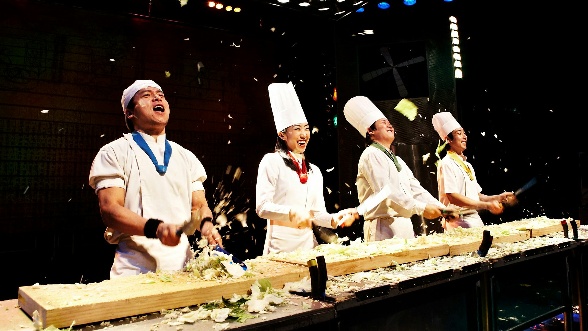 Four chefs wildly chop vegetables as pieces fly through the air.