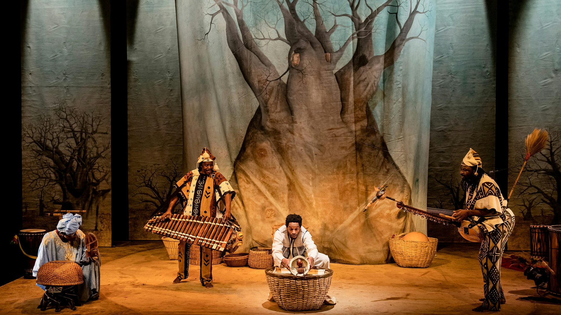 A bafalon player and kamele n'goni player stand to the left and right of a man holding a tiny puppet of a boy. Behind them is a backdrop of a baobab tree.