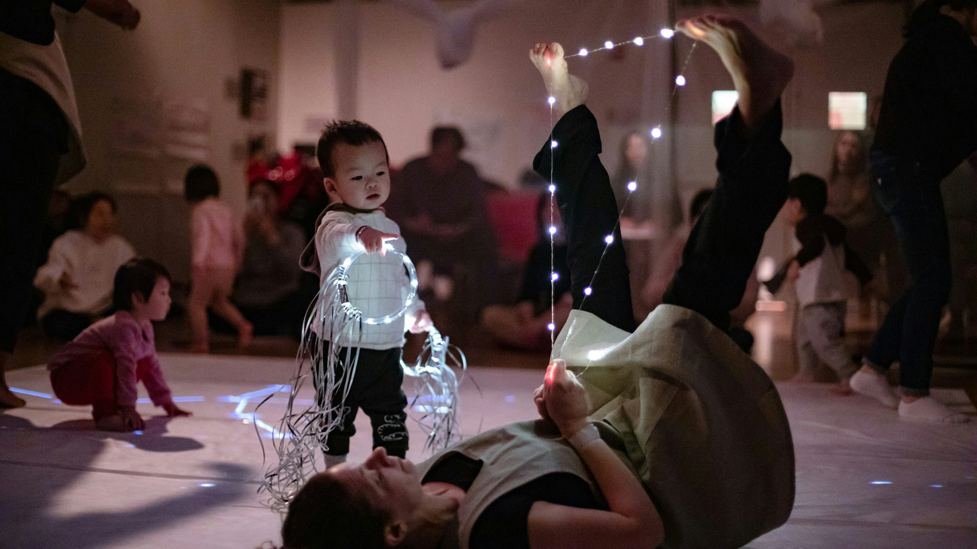 A baby points at an actor laying on the floor, holding string lights in between her feet.