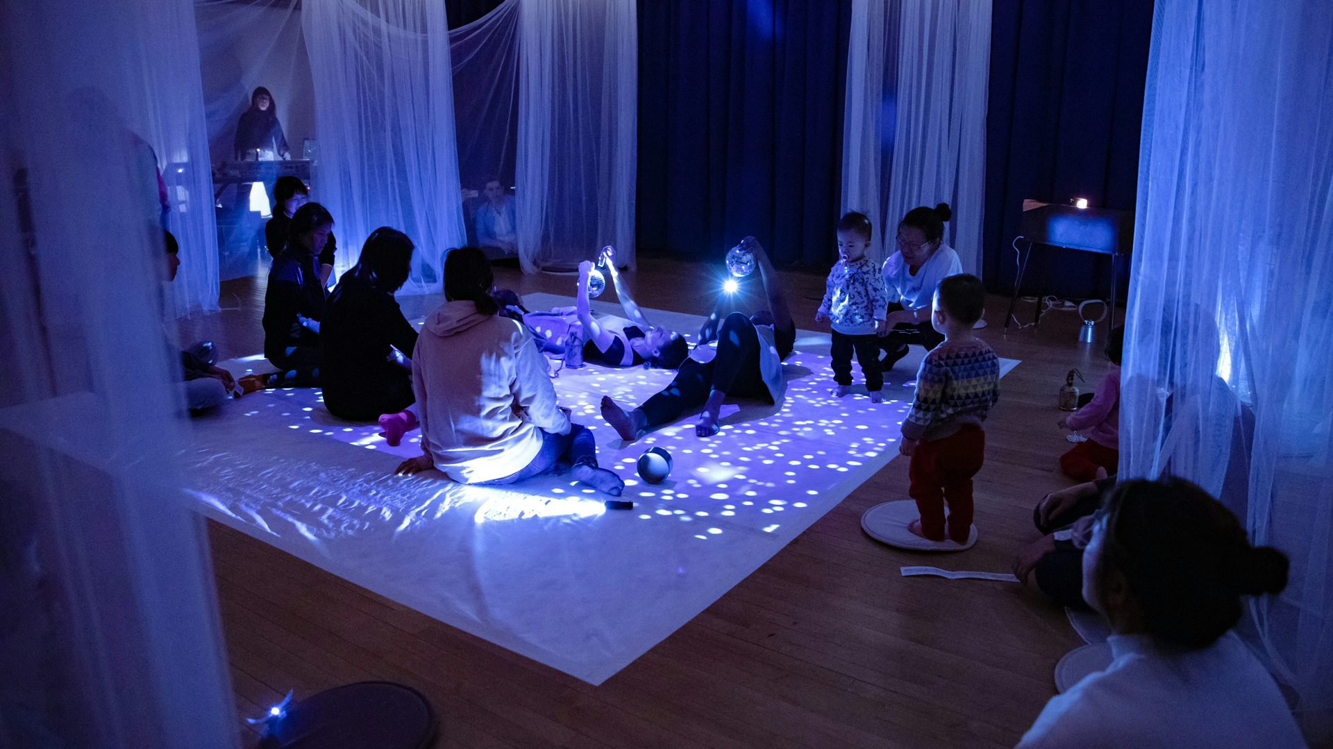 Audience members sit on the floor of a large studio that is illuminated by light reflections from disco balls.