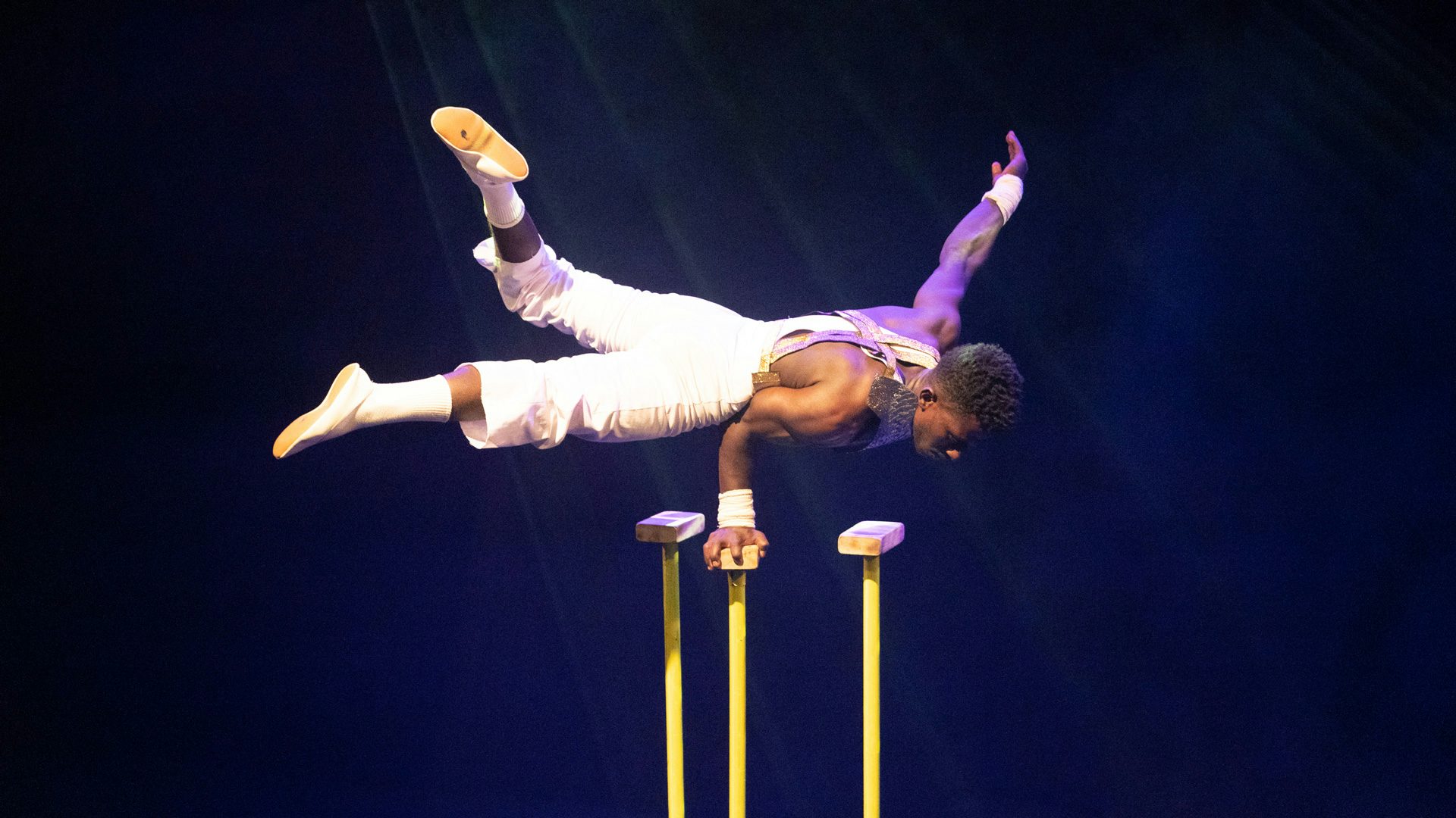 A shirtless acrobat in overalls hand balances on stilts.