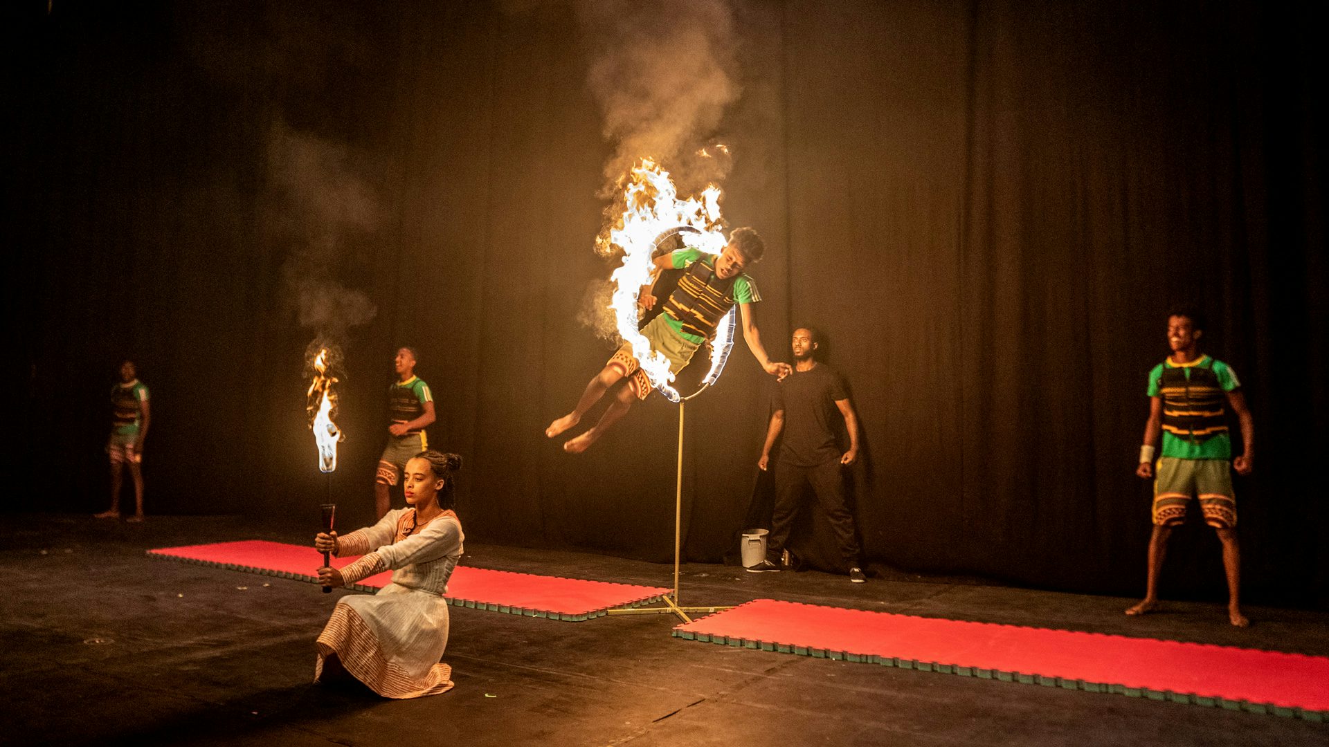 A teenage acrobat jumps through a hoop that's on fire, as other acrobats stand behind him.