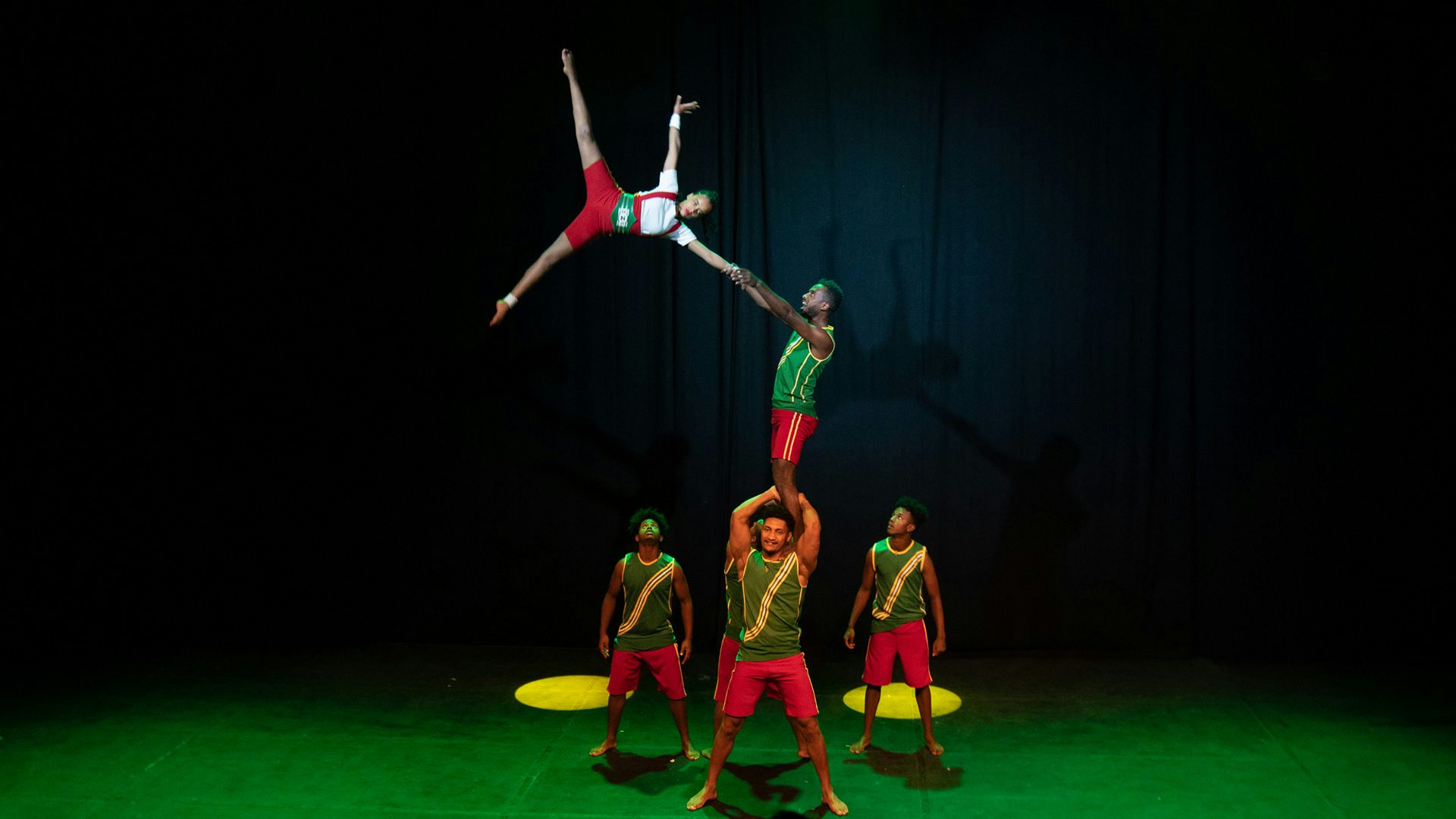 Holding onto one another by the wrists, a man suspends a woman in the air, while balancing on the shoulders of two other acrobats.