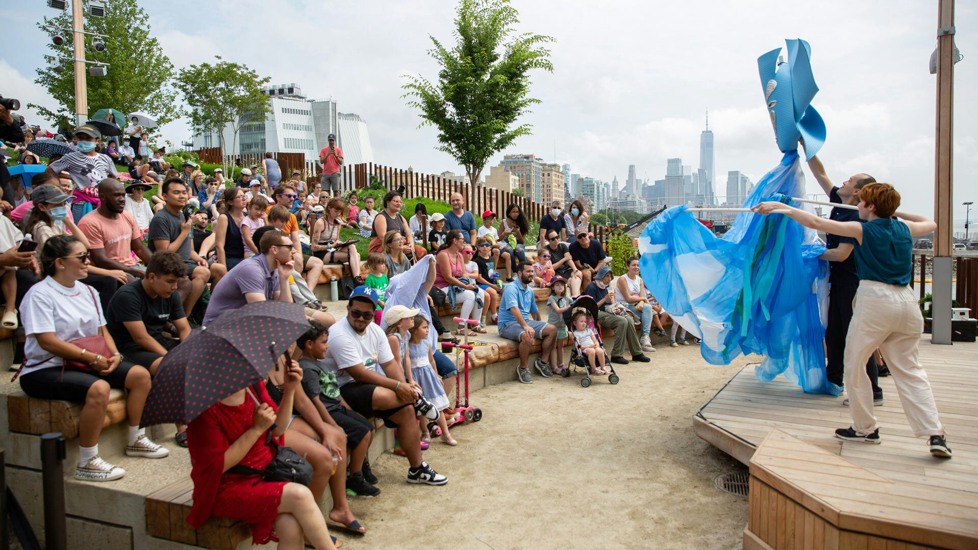 A crowd of people in an outdoor auditorium watch artists perform with a whale puppet onstage.