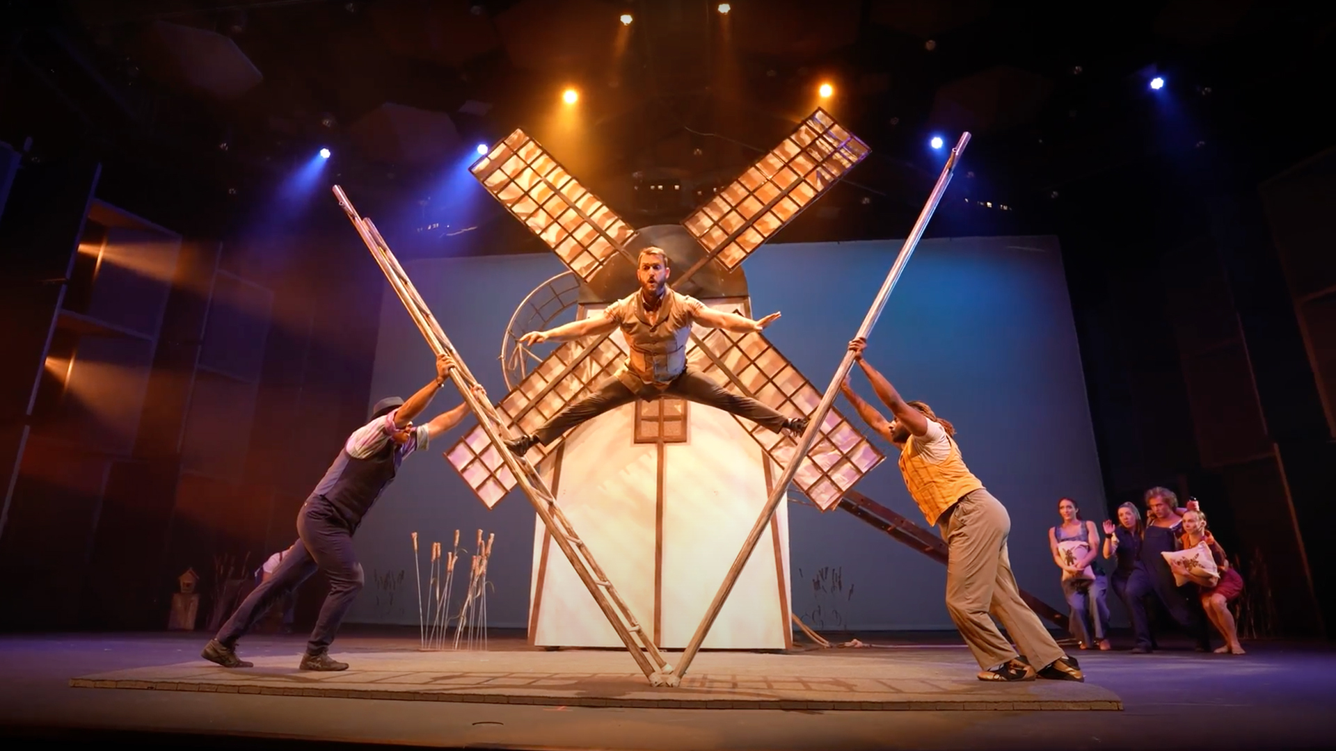 Photo of a performer standing on two falling ladders in the shape of a V. Behind is a windmill.