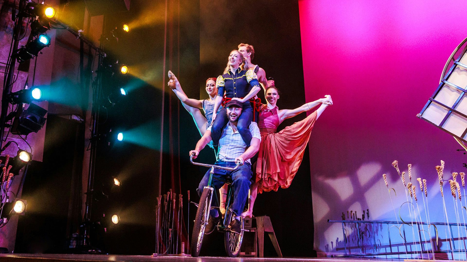 Five people balancing on a moving bicycle on stage.