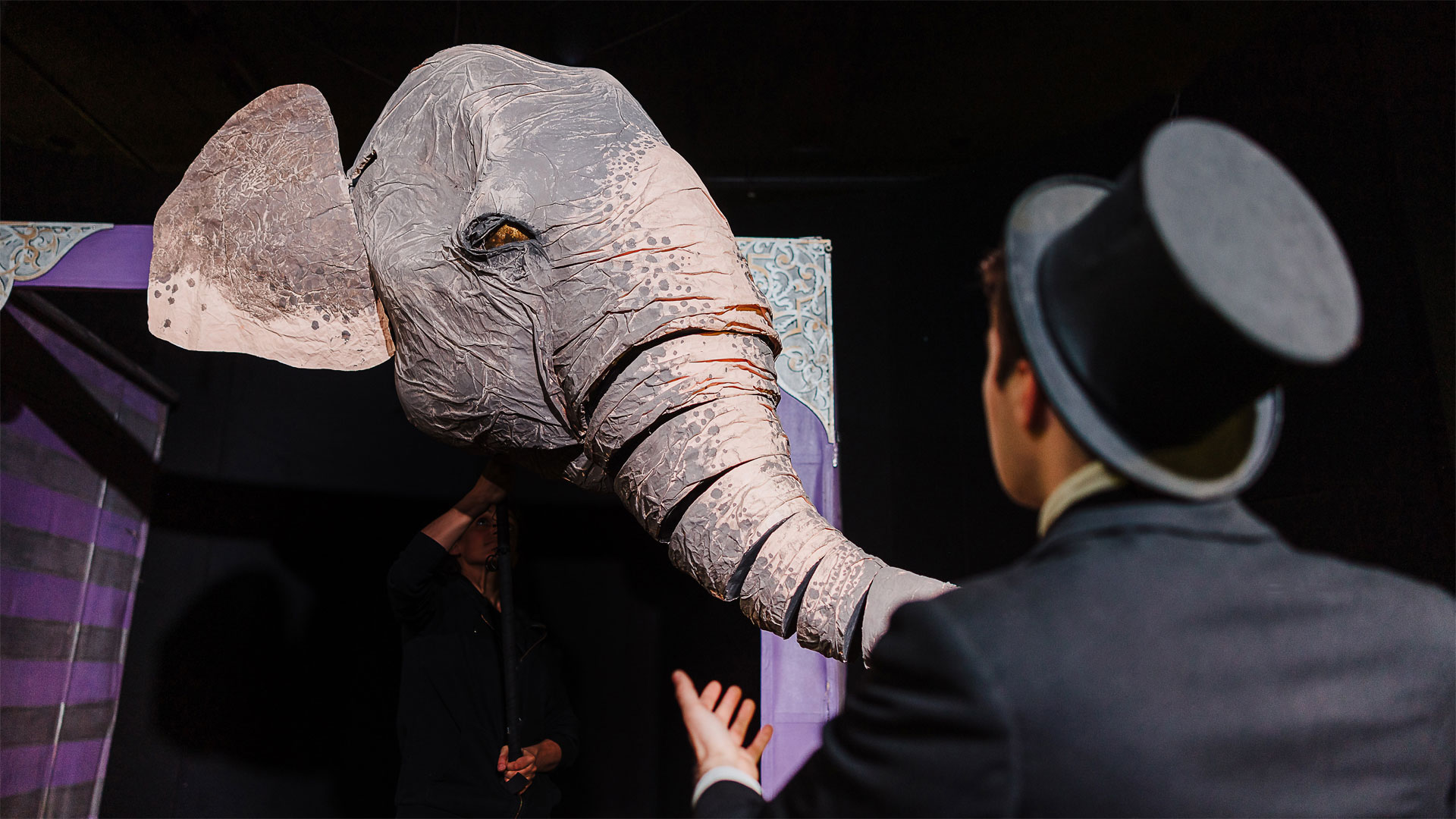 Puppet of an elephant head facing a person wearing a suit and top hat shown from behind