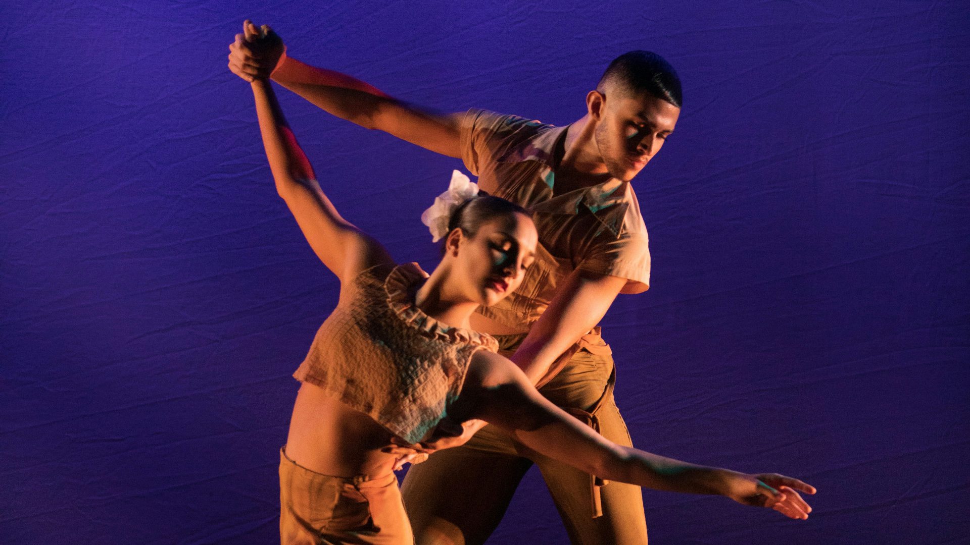 Two people in tan clothes holding each other and looking down and to the right