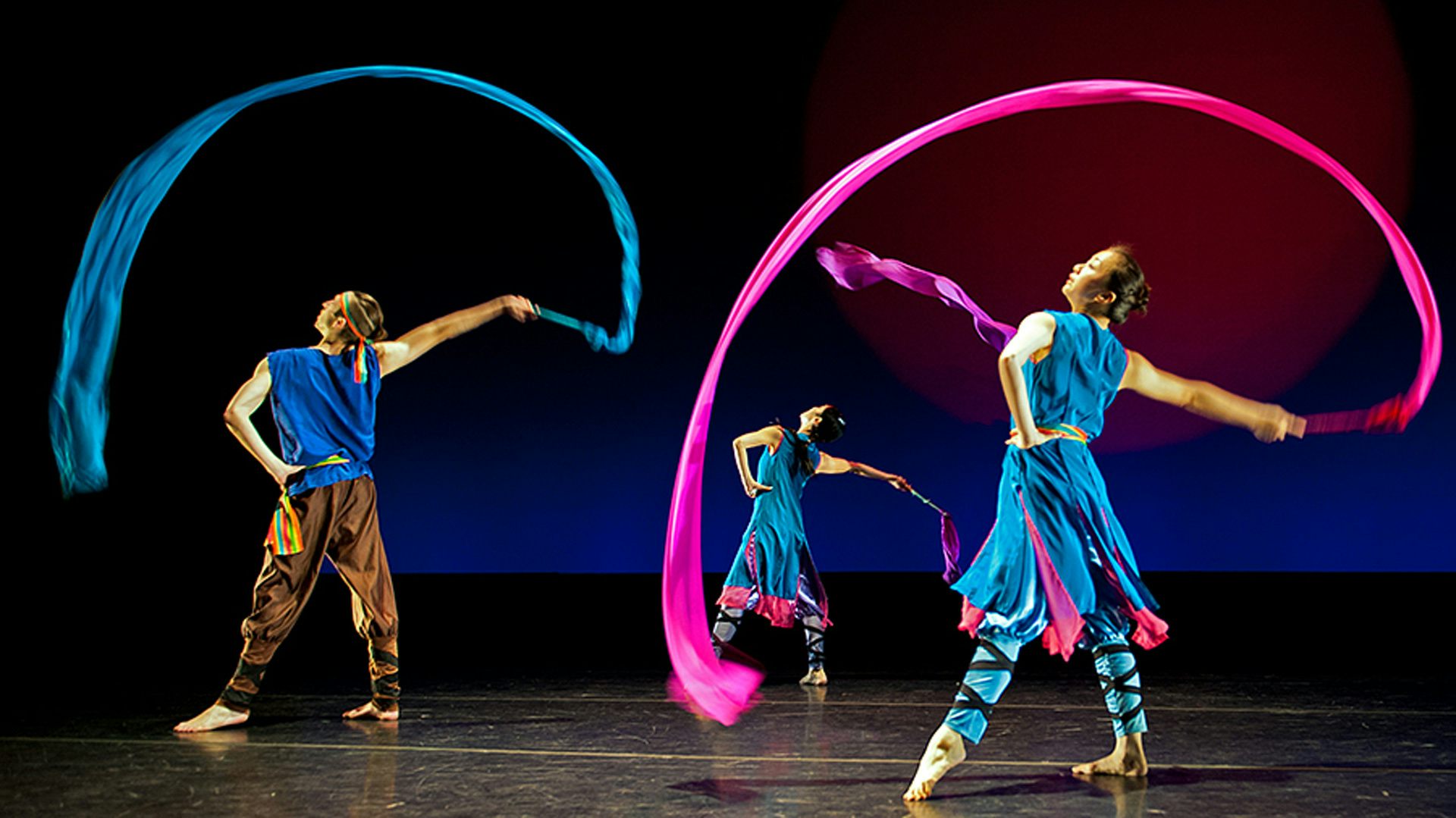 Three dancers wearing blue clothes looking to the left with one hand on their hips and the other hand holding a blue or pink ribbon that's arching over them