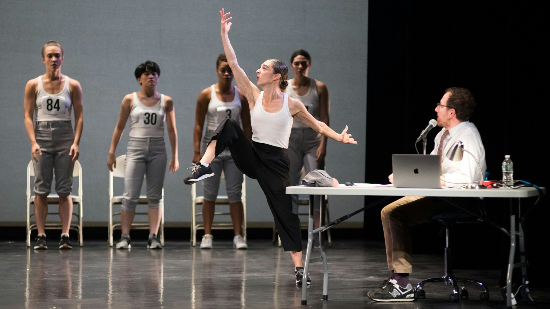 A person dancing with one arm and leg in the air while four dancers with numbers on their chests and a person sitting at a table with a computer look on