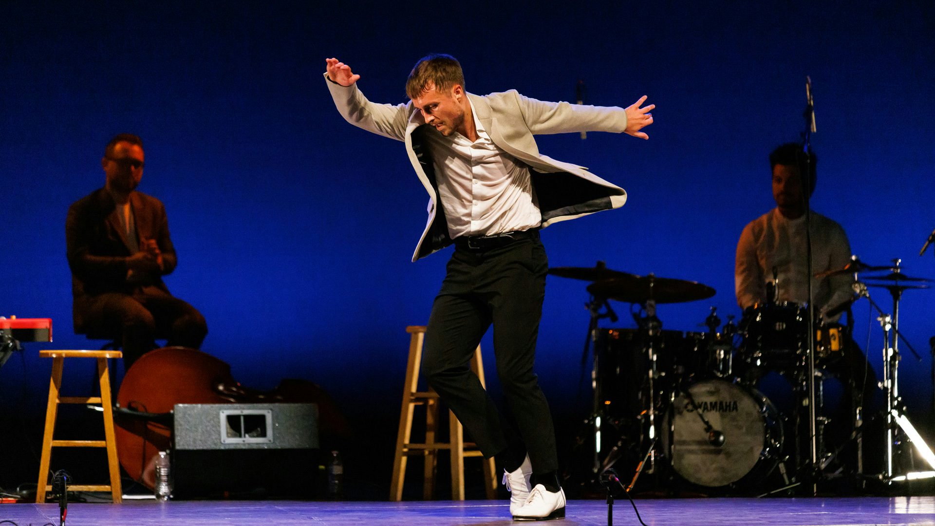 A person in a white shirt and jacket and black pants looking down with their arms up while standing in front of a two-person band