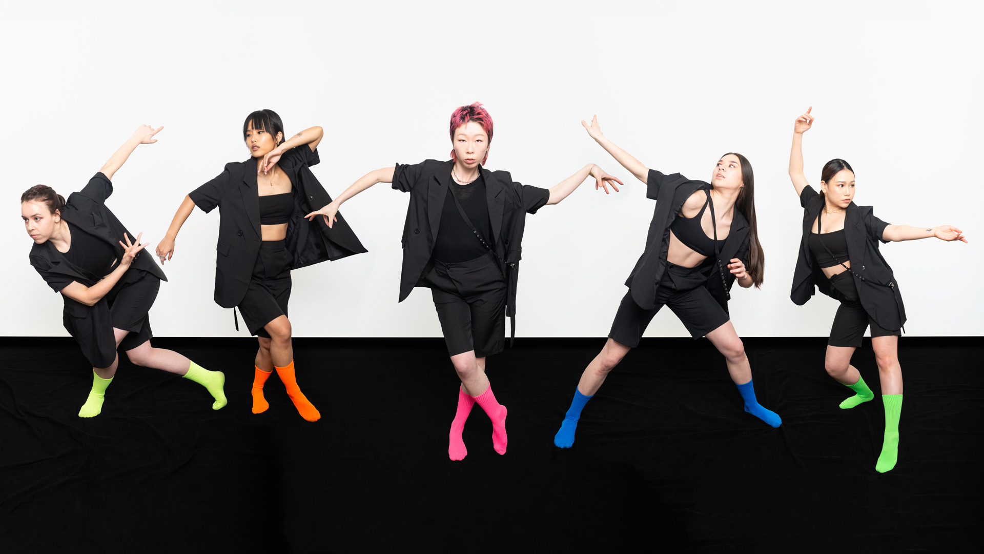 Five dancers posing on a black floor in front of a white background while wearing black clothes and different brightly-colored socks