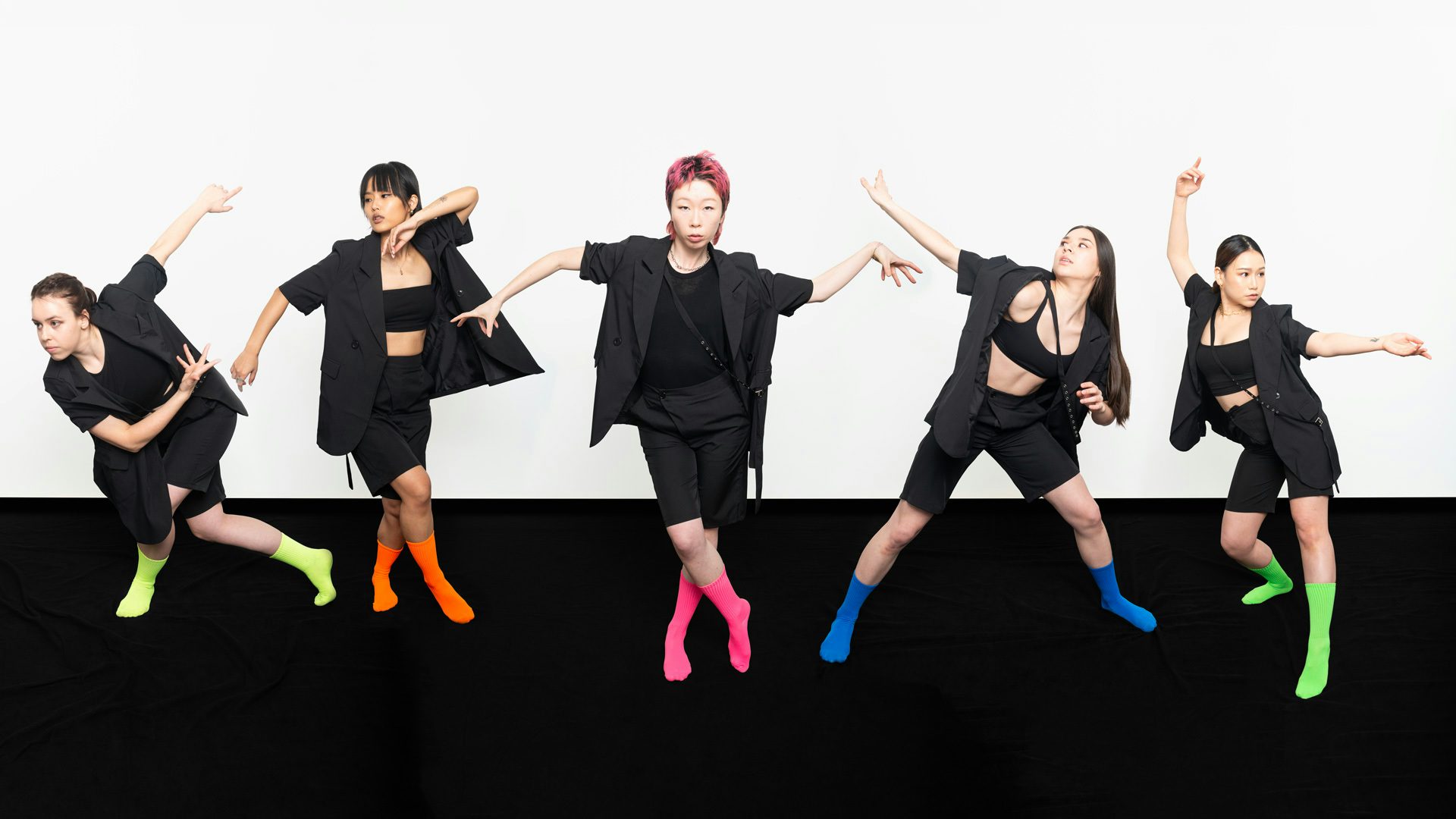 Five dancers posing on a black floor in front of a white background while wearing black clothes and different brightly-colored socks
