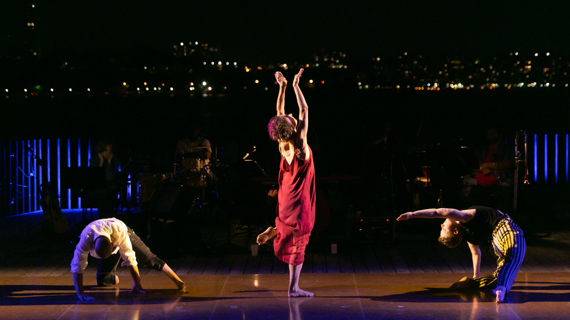 A person in a long, red dress facing the back while standing on one leg and two people on the ground on either side of them
