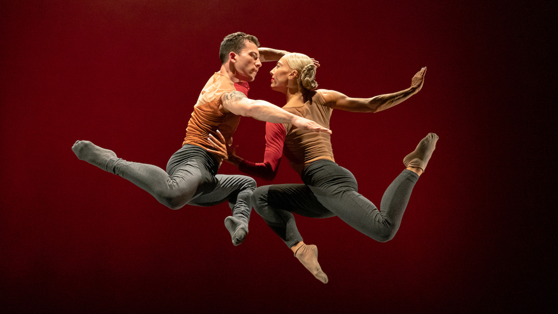 Two people jumping in the air and holding onto one another against a dark red background