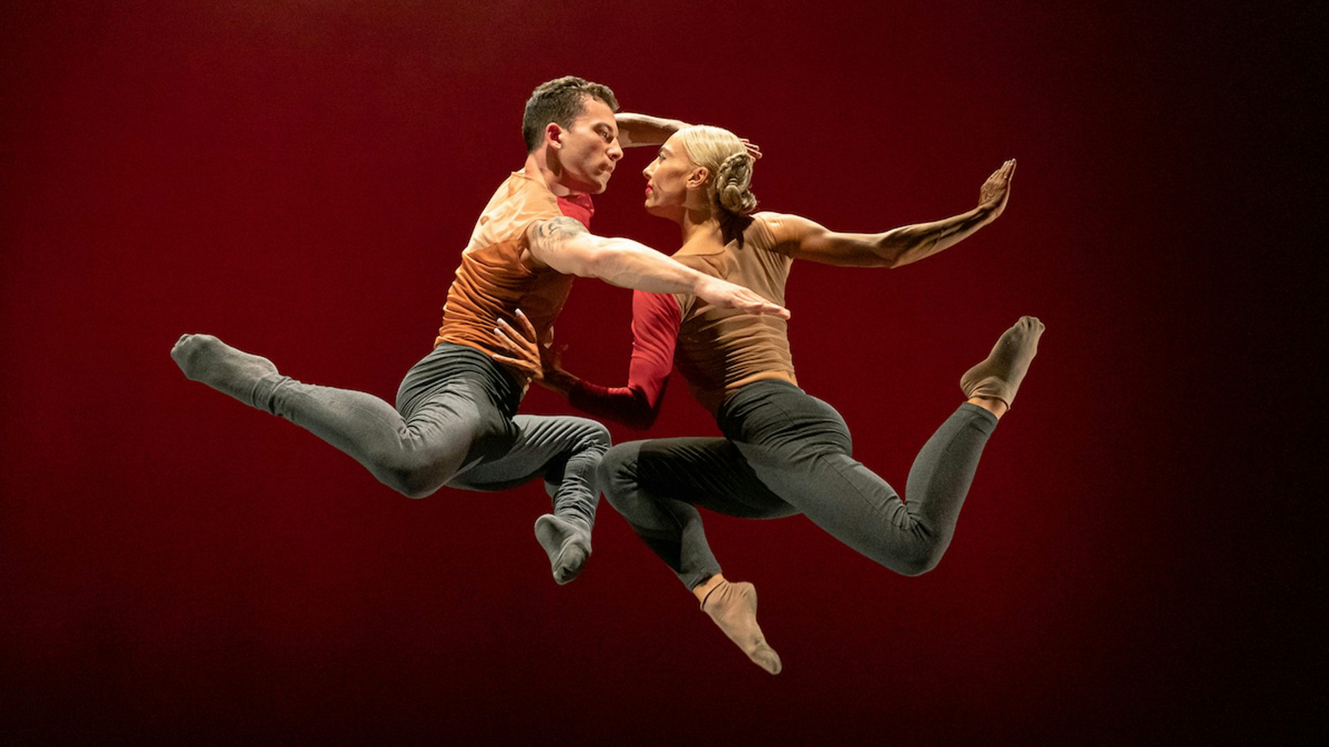 Two people jumping in the air and holding onto one another against a dark red background
