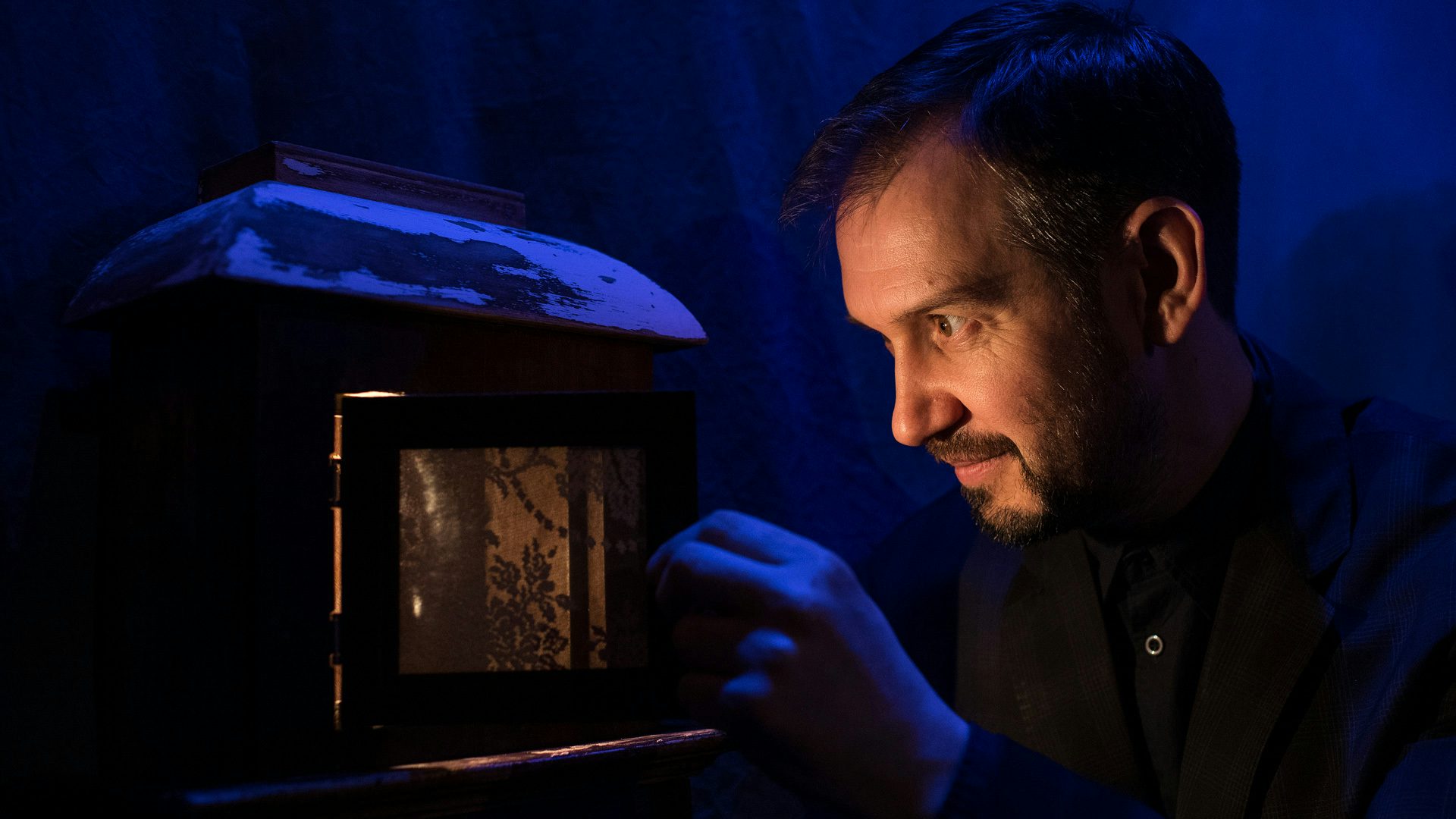 A man with a beard looks excitedly into a small wooden box with a warm orange/yellow light coming out from it.