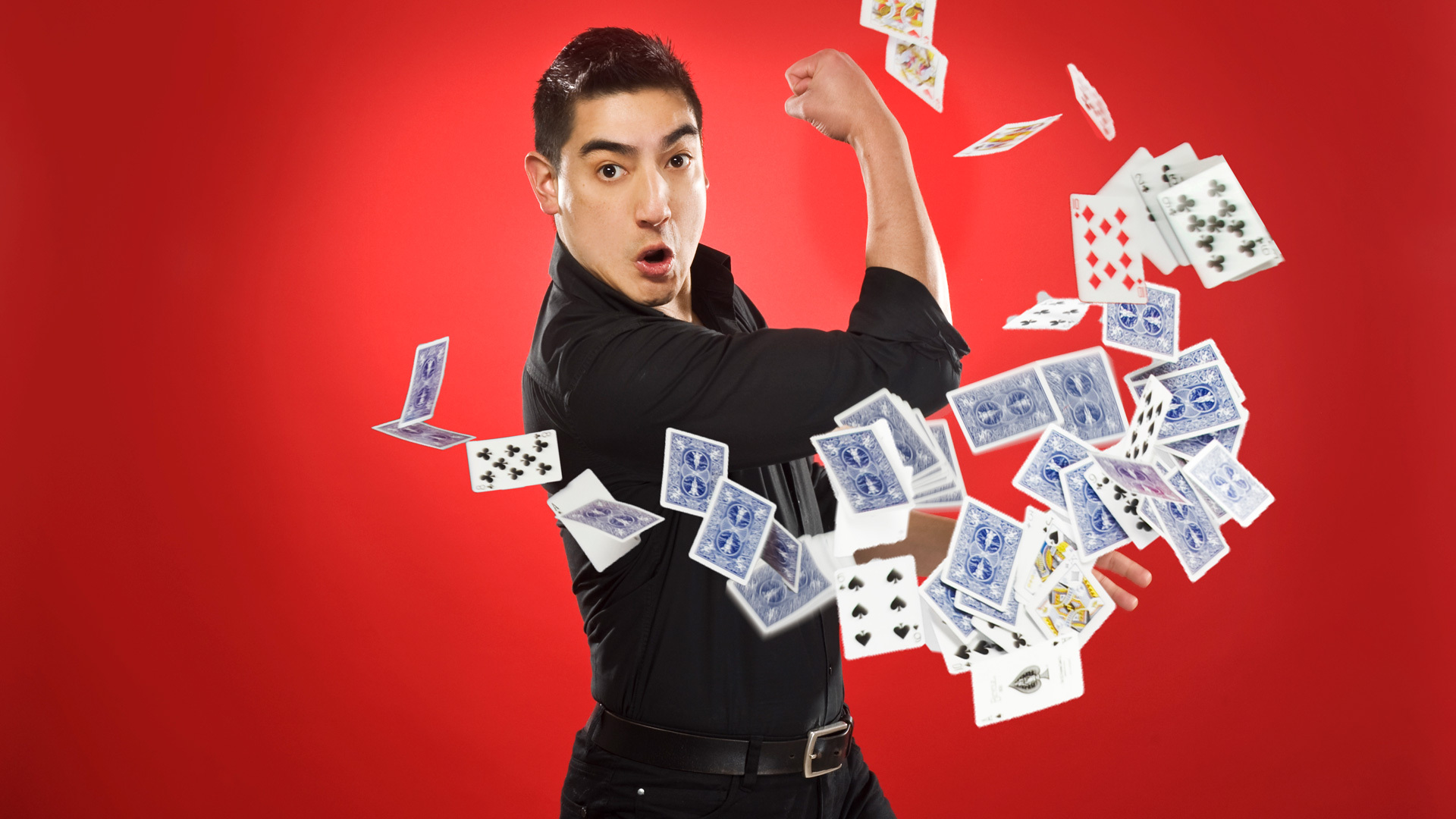 A man wearing a black shirt throws playing cards in a flourish against a red background.