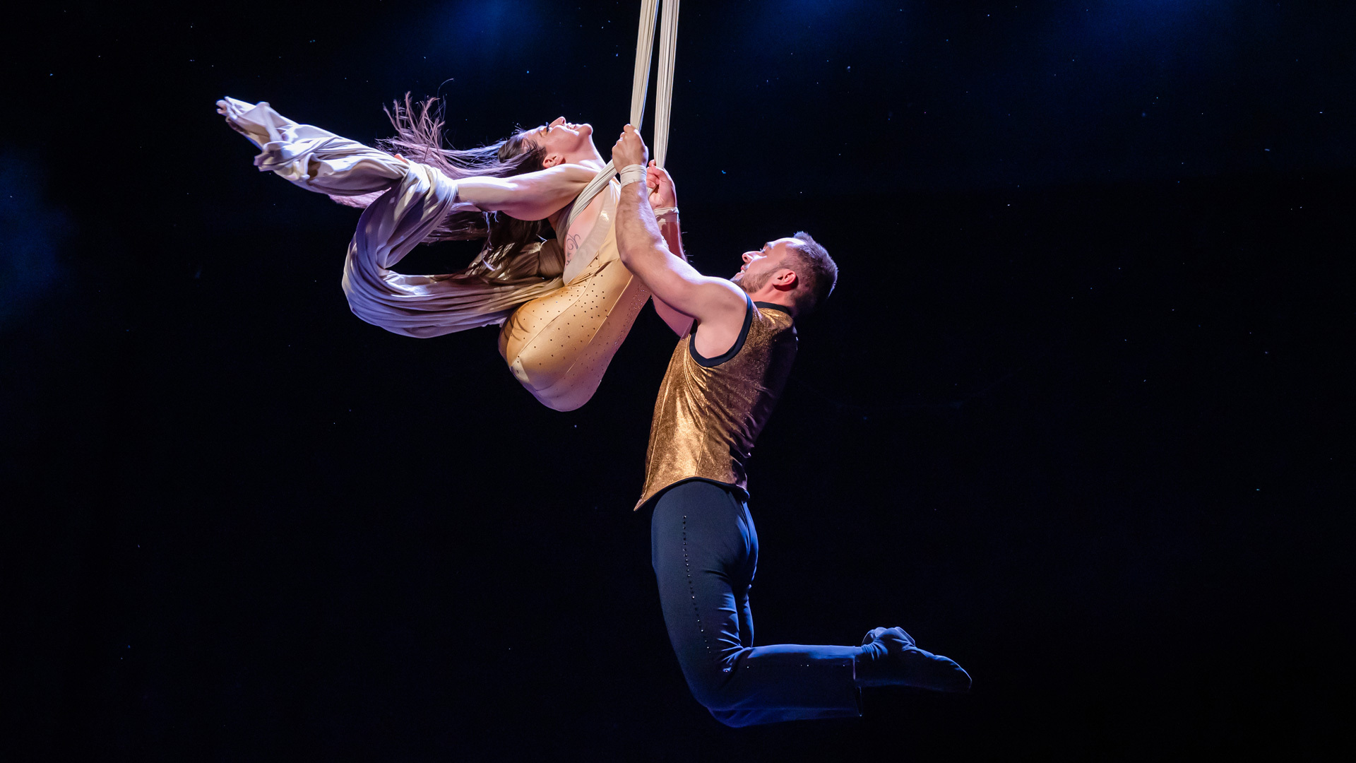 A man and a woman wearing gold outfits perform aerial silks.