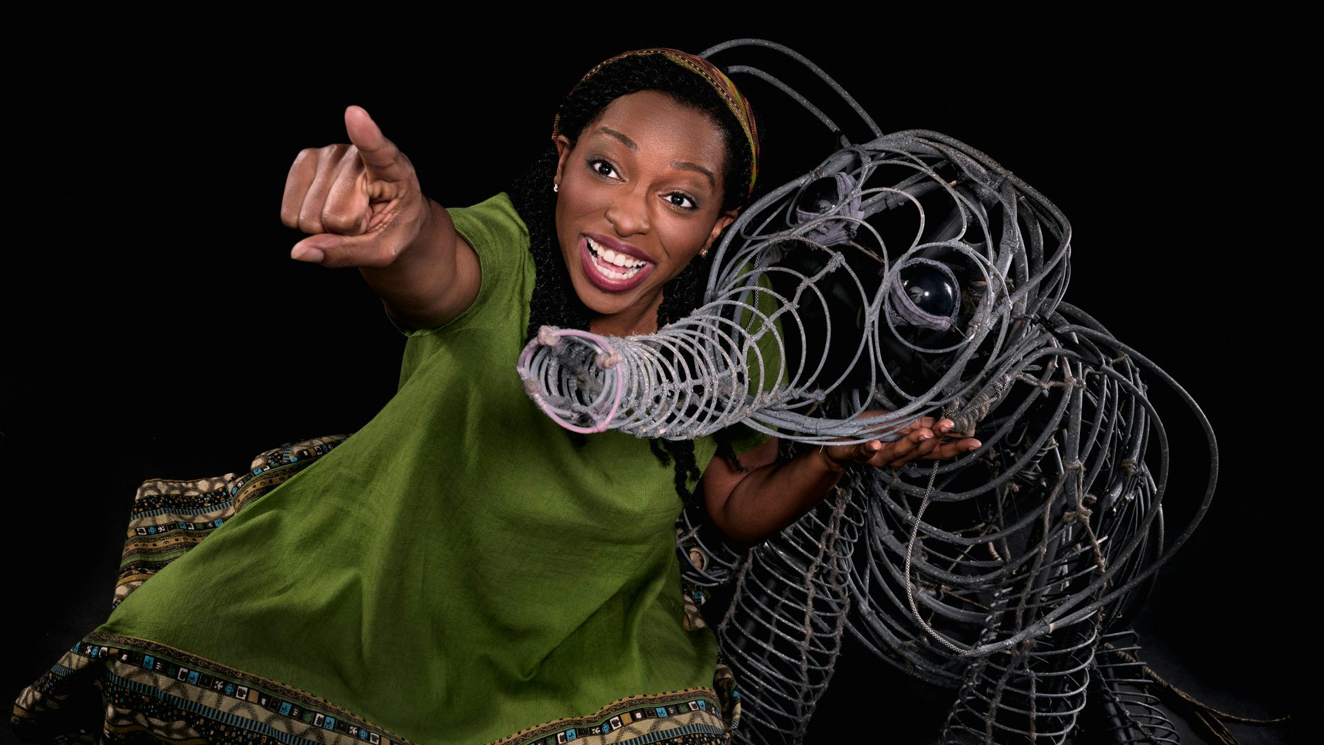 A woman in a green dress holds a wire sculpture/puppet shaped like a small elephant and points off to the distance to the left.