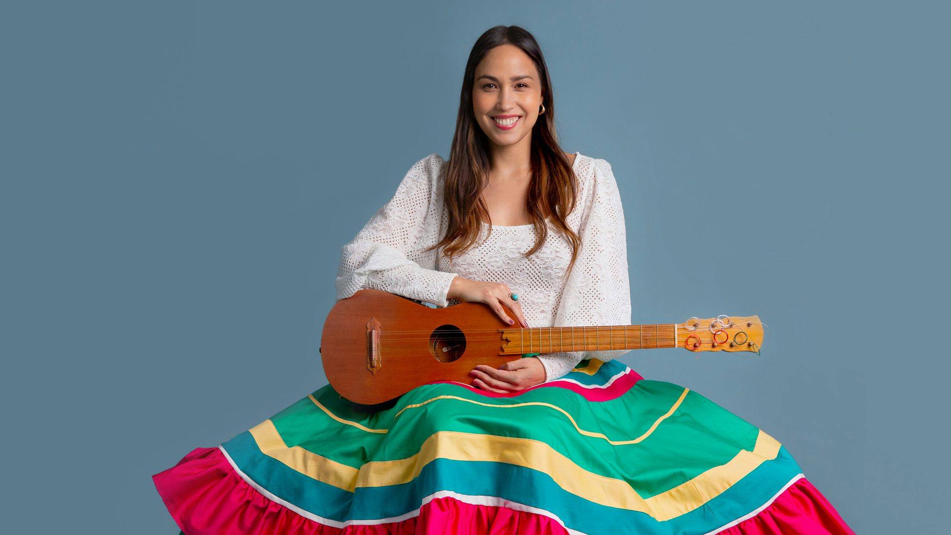 A woman in a colorful skirt smiles and sits against a blue background while holding a jarana—a string instrument resembling a guitar with Mexican origins.