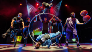 A group of seven people on a stage perform stunts with big metal hoops, bicycles and basketballs.