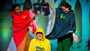 A person wearing a red raincoat and a person wearing a navy raincoat pour water onto a man in a yellow raincoat's head from blue and purple watering cans.