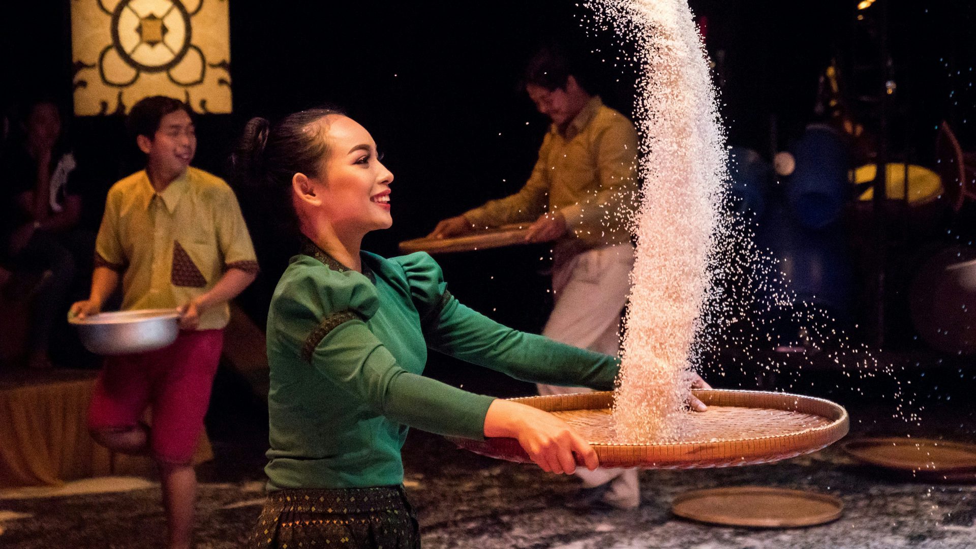A woman wearing a green shirt faces the right and throws rice from a tray into the air while two men hold wooden bowls in the background.