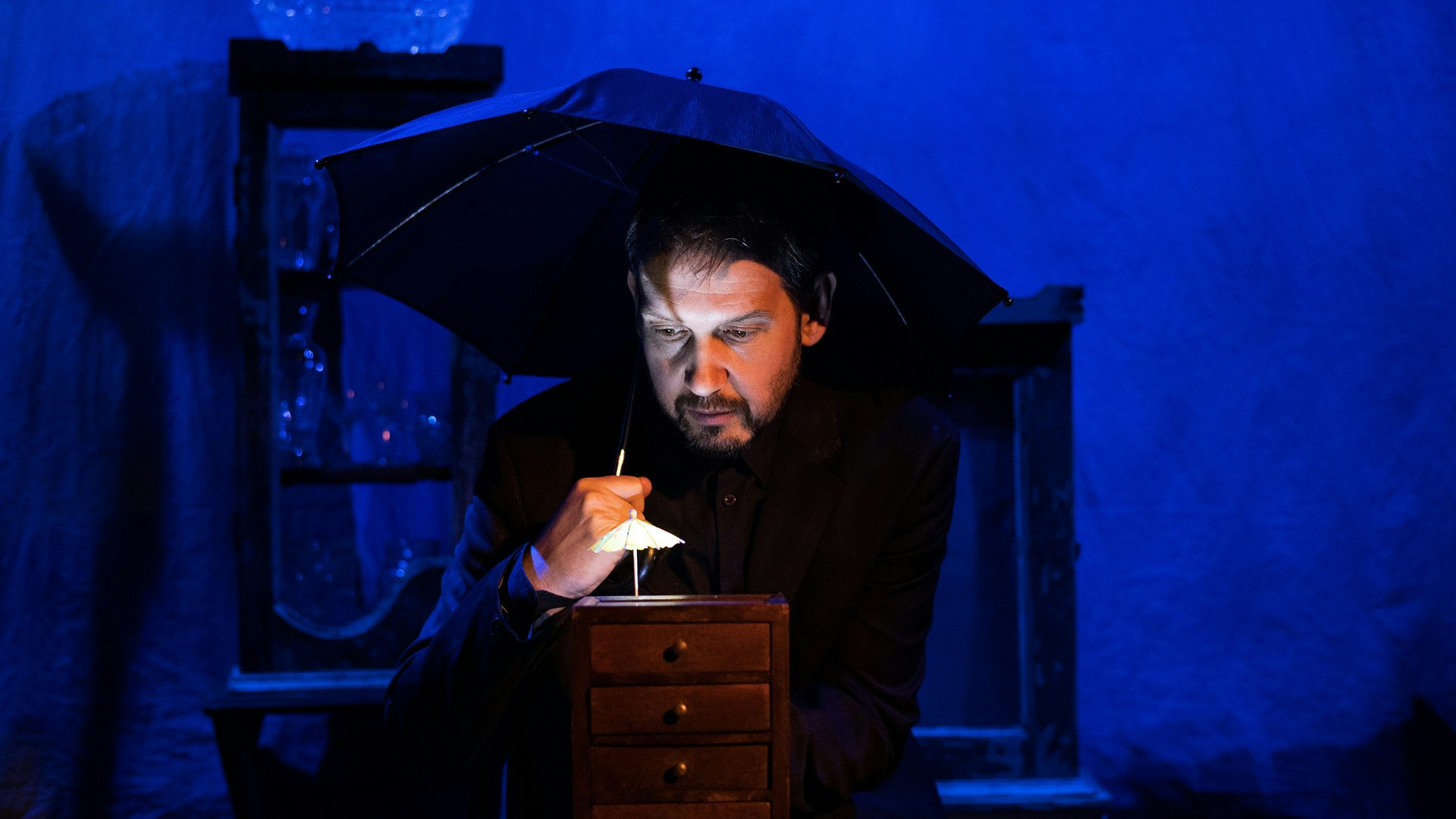 A man with a beard holds a tiny cocktail umbrella with his fingers over a tiny wooden chest. The man also holds a normal-sized umbrella while being backlit by blue light. The tiny cocktail umbrella is illuminated underneath by a small white light.