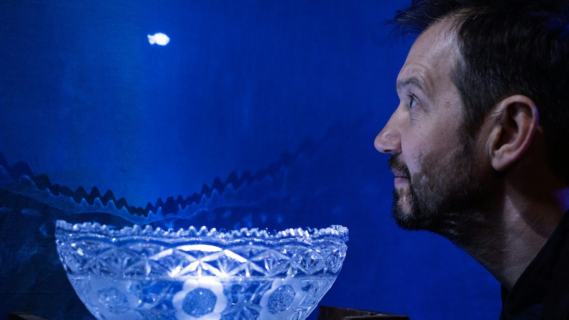 A man with a beard looks at a small white silhouette of a fish over a large, ornate, glass bowl.