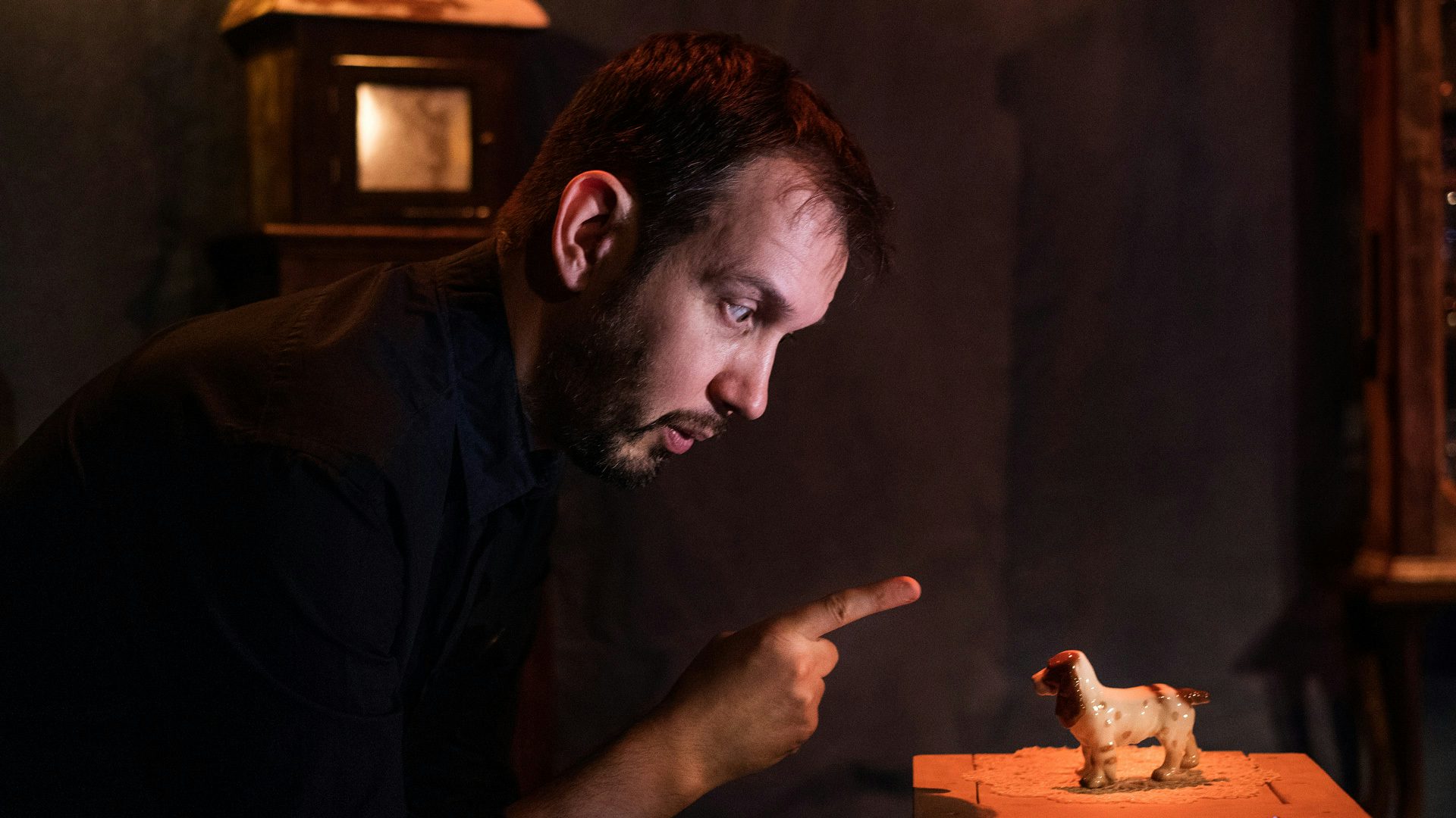 A man with a beard points at a toy dog on a small table as if reprimanding it. Warm, orange lights illuminate the stage.